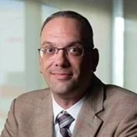 Close-up photo of a man smiling, wearing black glasses and a green shirt, in an indoor setting with bright lighting.