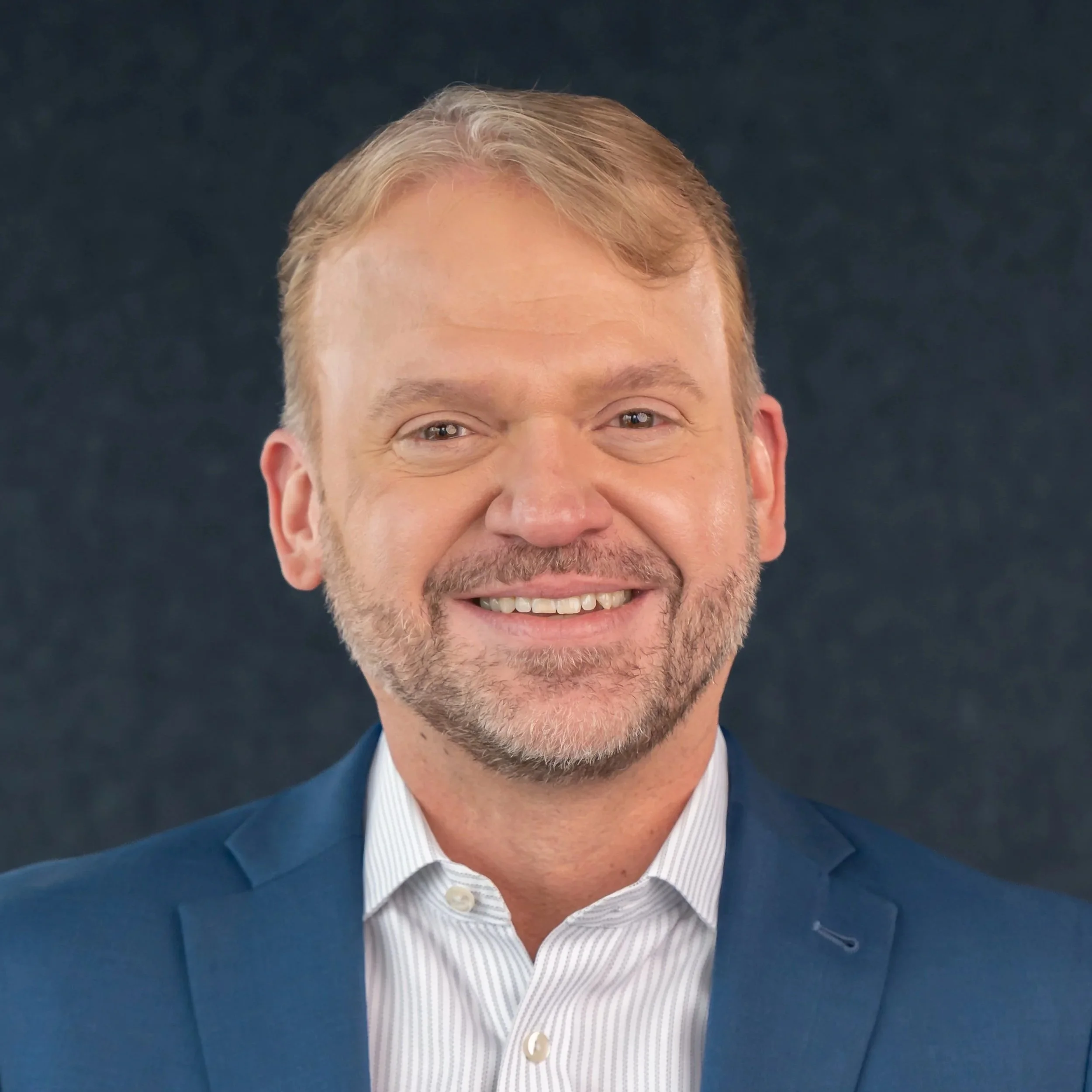 A man with short light brown hair, a beard, wearing a dark suit, light pink shirt, and light gray tie, smiling against a plain gray background.