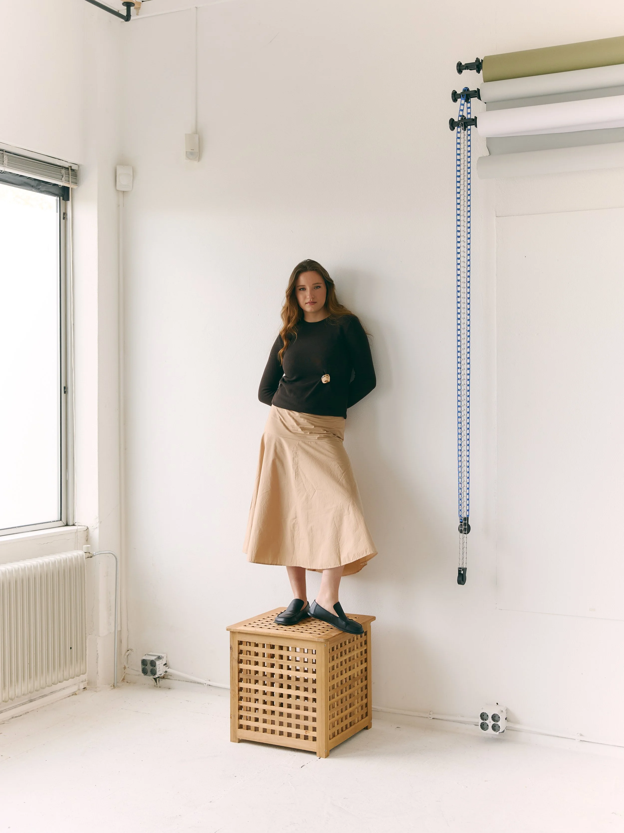 A woman with long hair standing on a wooden crate inside a white room. She is wearing a black top and a beige skirt, and has one hand in her pocket. There are rolls of paper or fabric hanging on the wall to her right, and a window with a radiator below it on her left.