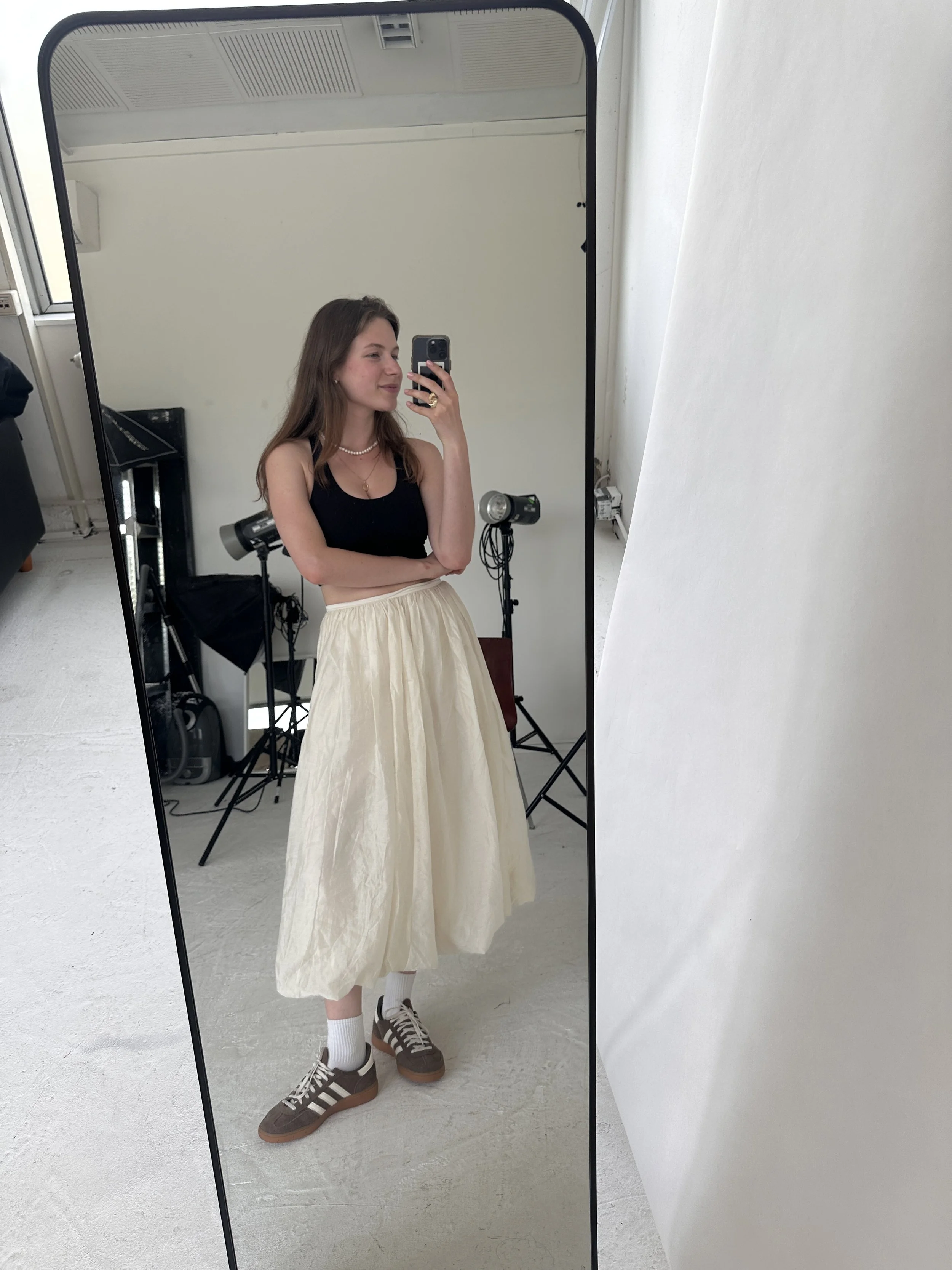 A young woman taking a mirror selfie in a photography studio. She is wearing a black sleeveless top, a cream-colored skirt, white socks, and sneakers. She has necklace jewelry and is smiling slightly.