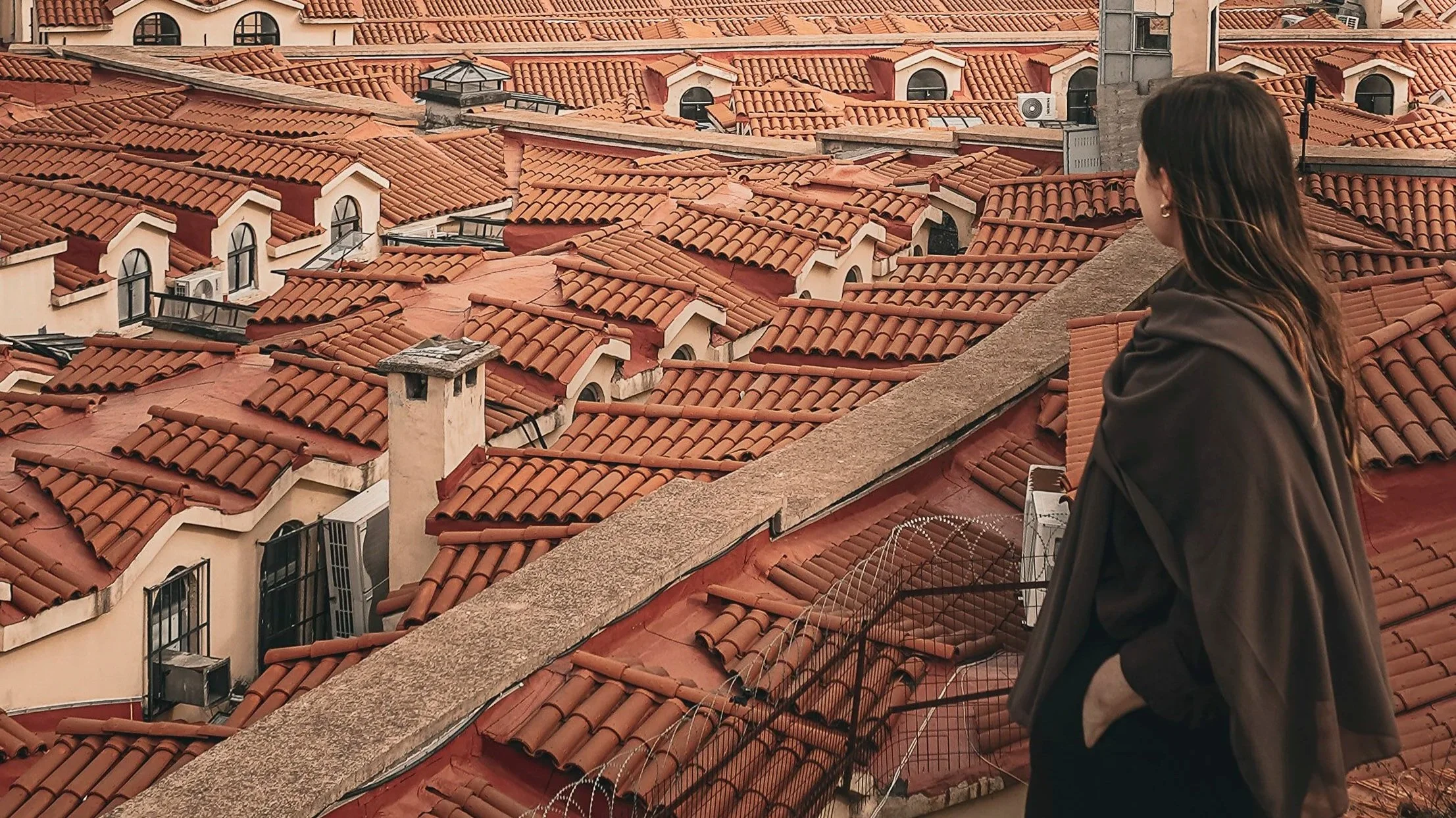 A woman with long brown hair wearing a dark jacket sits on a rooftop ledge overlooking a neighborhood of houses with red tiled roofs.