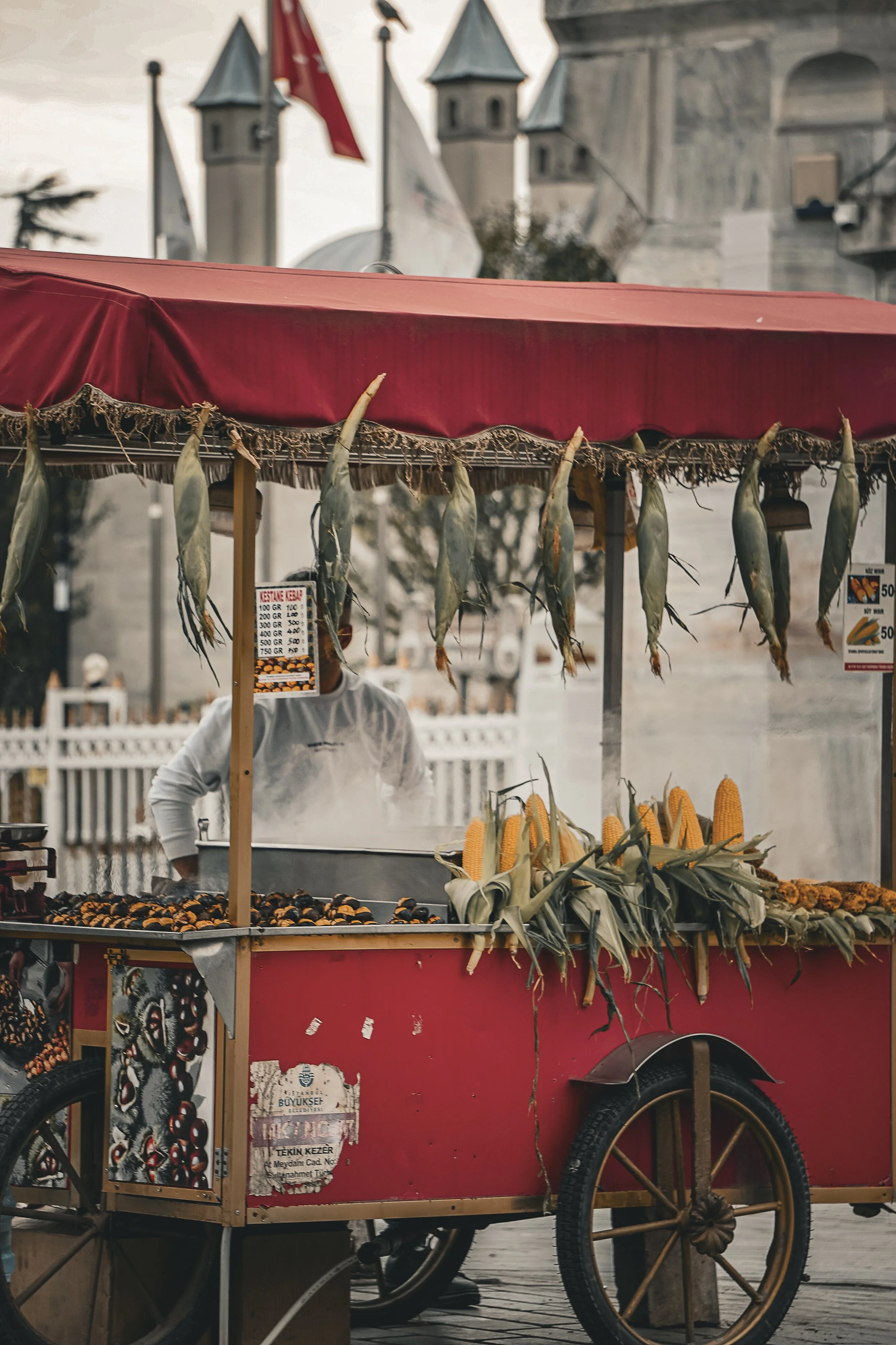 A red street food cart selling corn and other snacks, with a person behind it wearing a white shirt. The cart has a canopy with hanging fish and a menu sign with prices. In the background, there is a historic stone building with turrets, and flags flying.