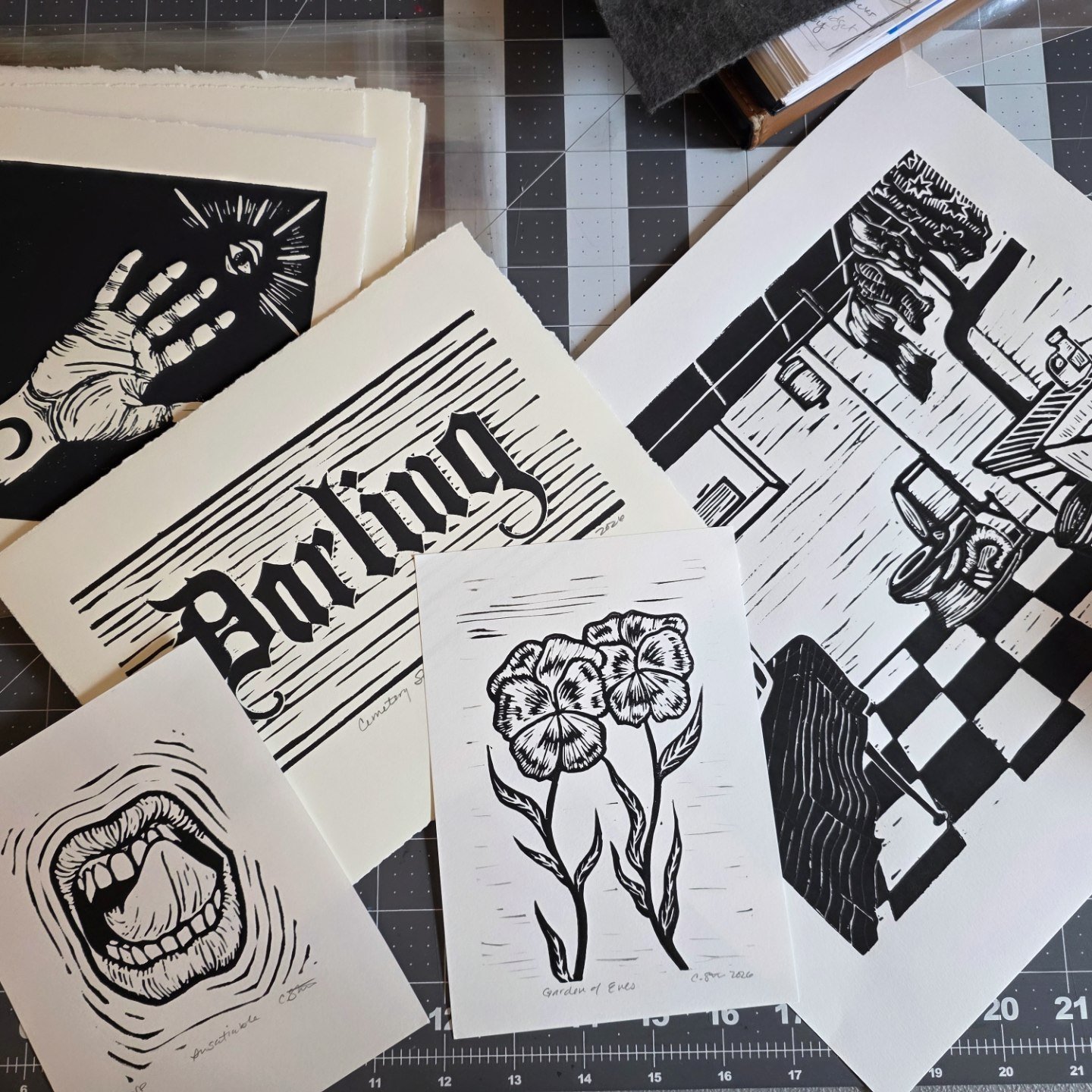 Black-and-white print artworks on paper, including a hand reaching out towards the third eye, the word 'Darling', lips, pansies, and a basement scene, spread out on a crafting mat.