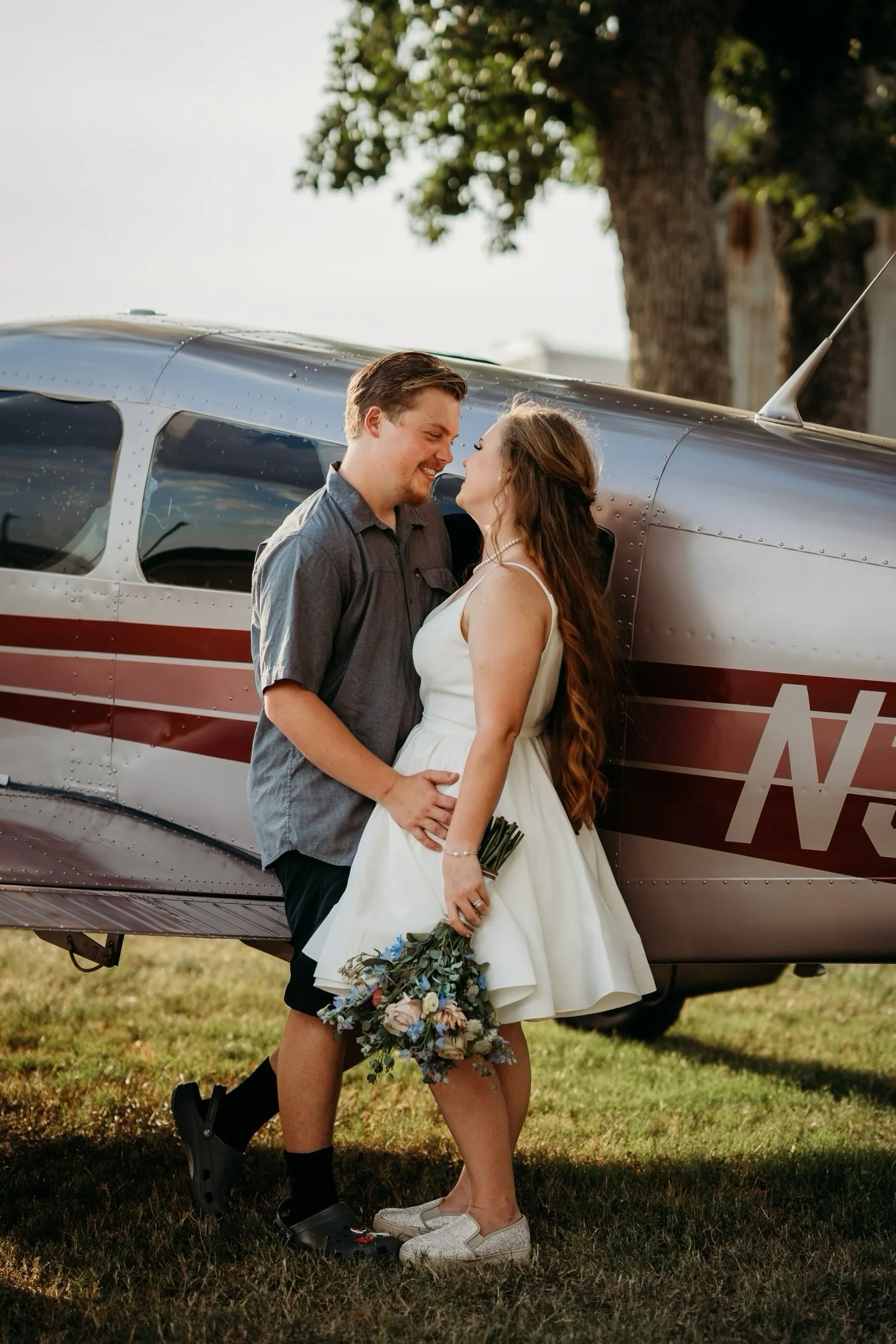 A couple stands close together outdoors, smiling at each other near a small airplane. The woman is holding a bouquet of flowers and is wearing a white dress and sneakers. The man is wearing a gray shirt and black shorts. A large tree is in the background, and the scene is lit by warm sunlight.