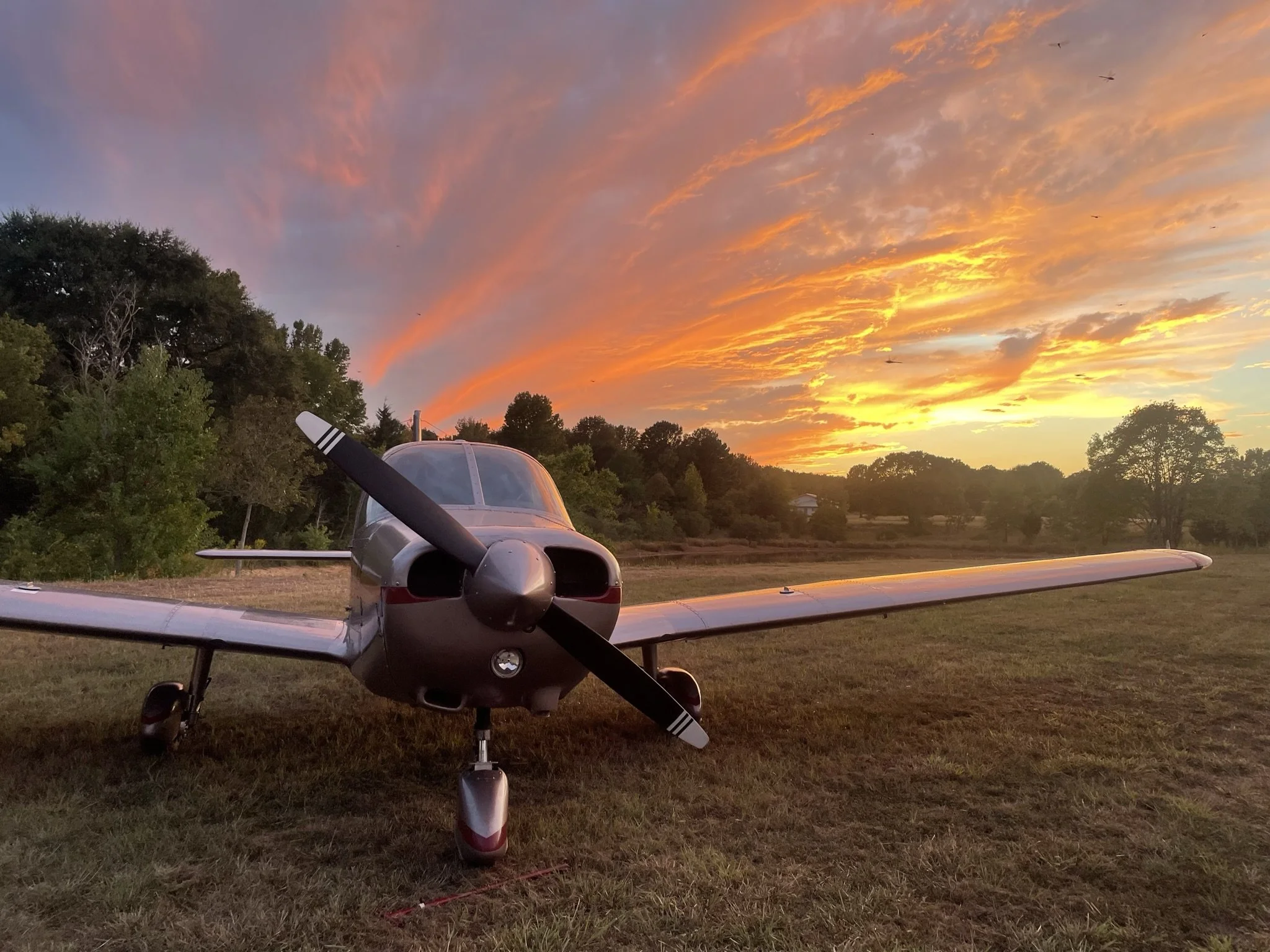 A small propeller airplane parked on a grassy field at sunset with a colorful sky filled with orange, pink, and purple clouds and distant trees.