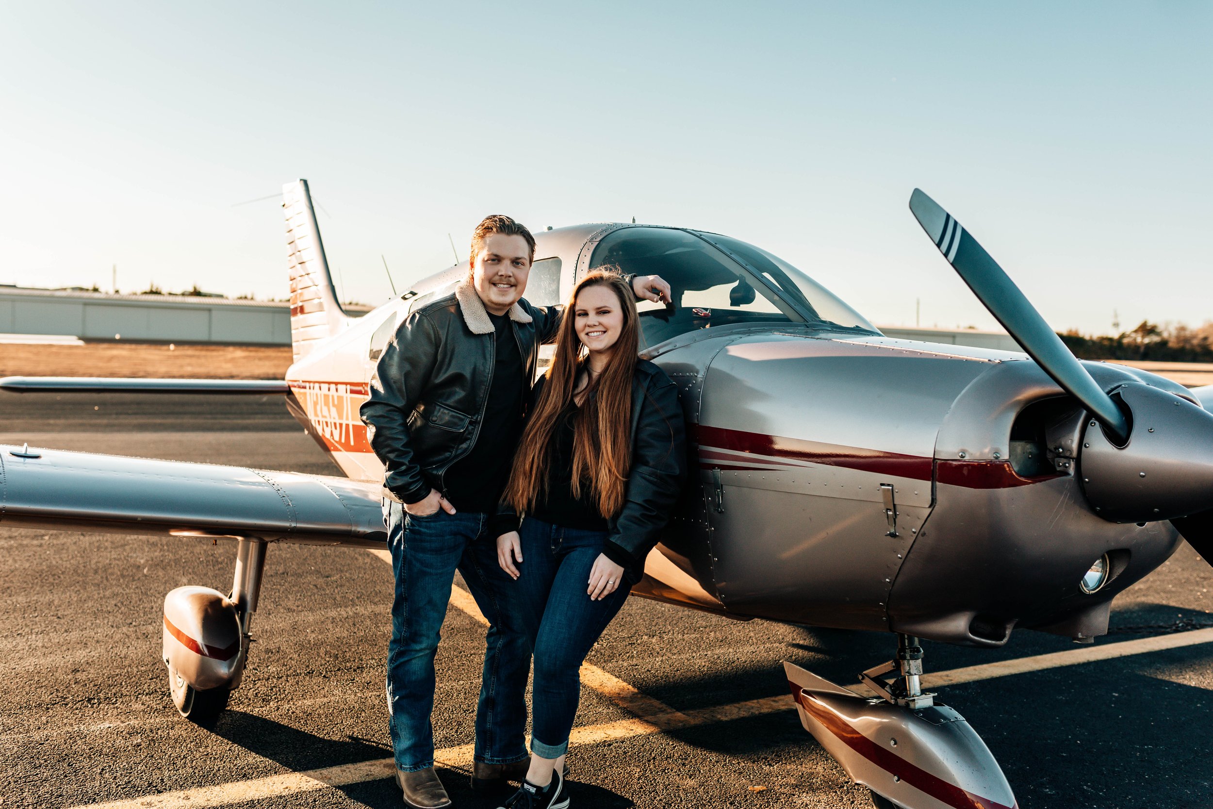 A young man and woman smiling and standing next to a small aircraft on an airport tarmac during sunset.