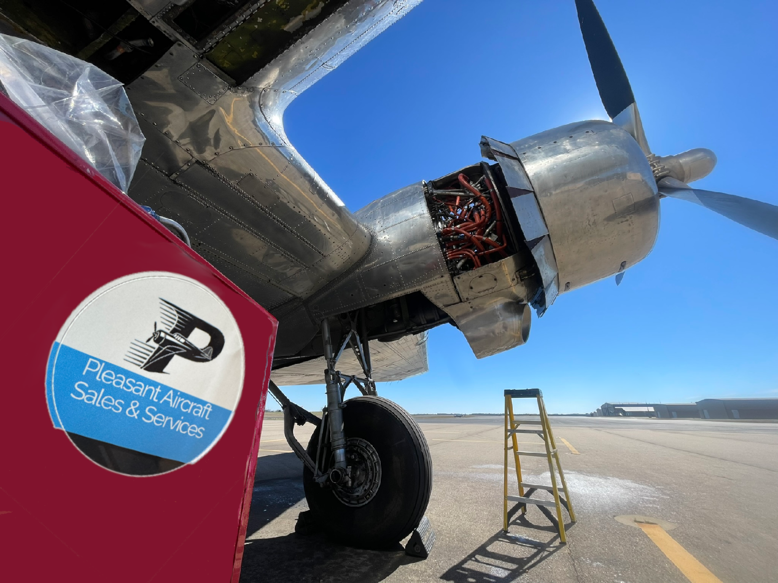 Close-up of an airplane's engine and landing gear on the tarmac, with a yellow stepladder nearby, under a clear blue sky. A red item with the Pleasant Aircraft Sales & Services logo is in the foreground.