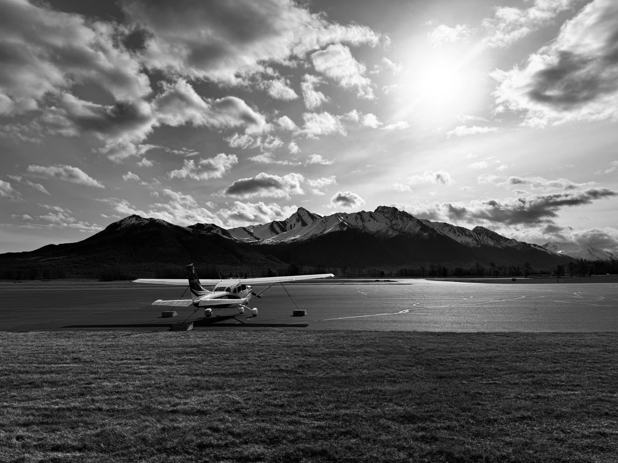 A small airplane parked on a tarmac with mountains and sky in the background, black and white