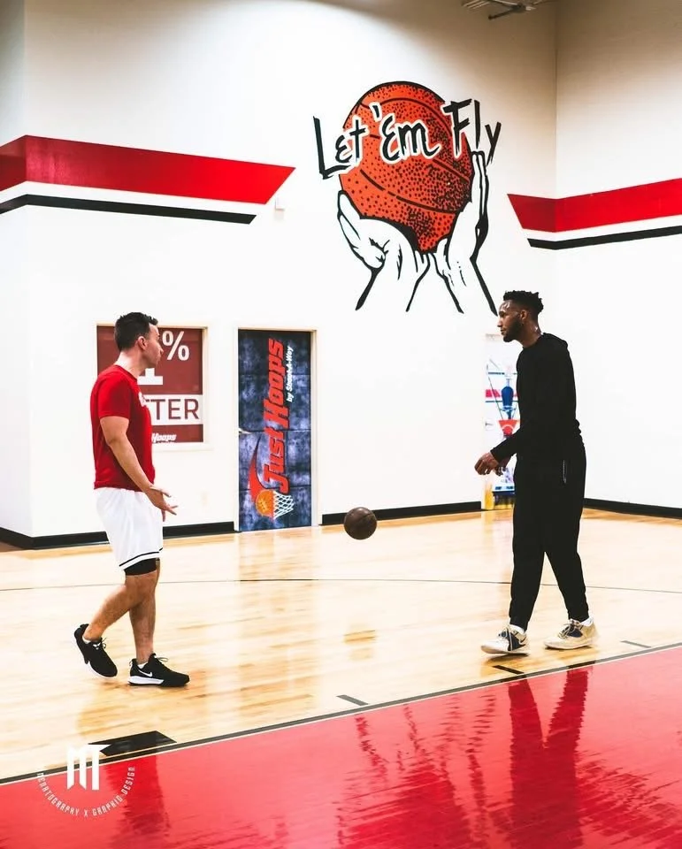 Two men playing basketball on an indoor court with a wall art that says "Let 'em Fly" and depicts a basketball above a pair of hands.