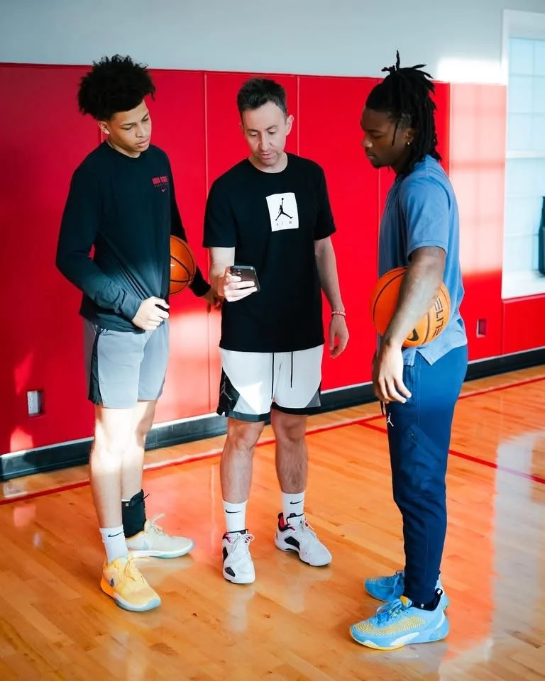 Two young men in basketball attire hold basketballs while listening to a coach or trainer showing information on a phone in a gym with red walls.