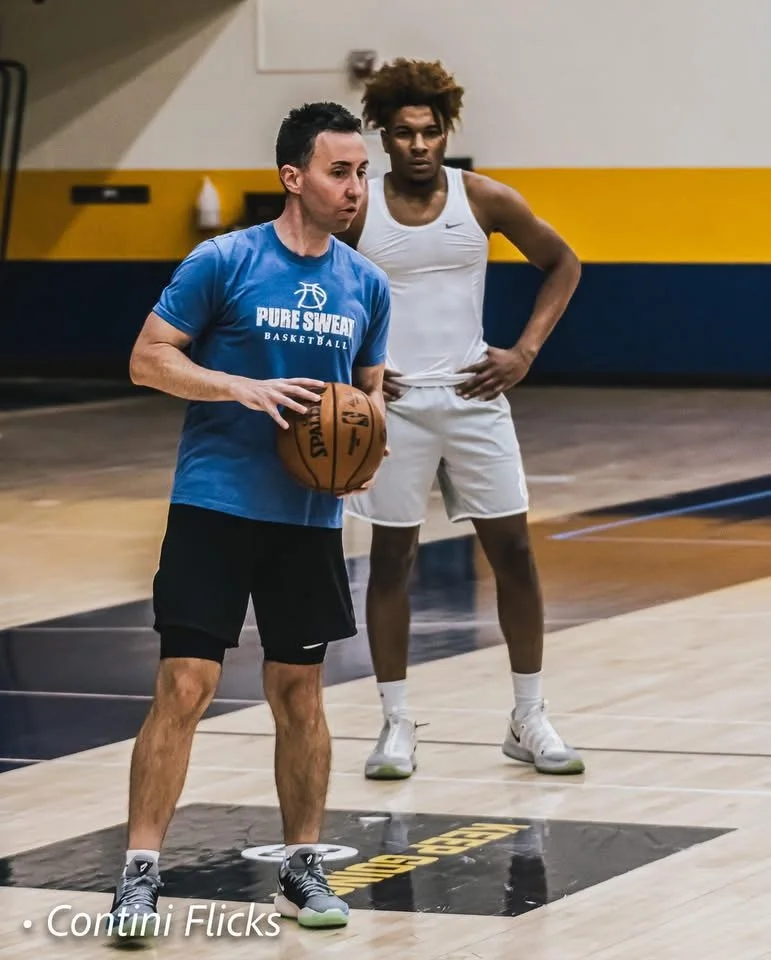 A coach in a blue t-shirt holding a basketball and a player in a white athletic outfit on a basketball court.
