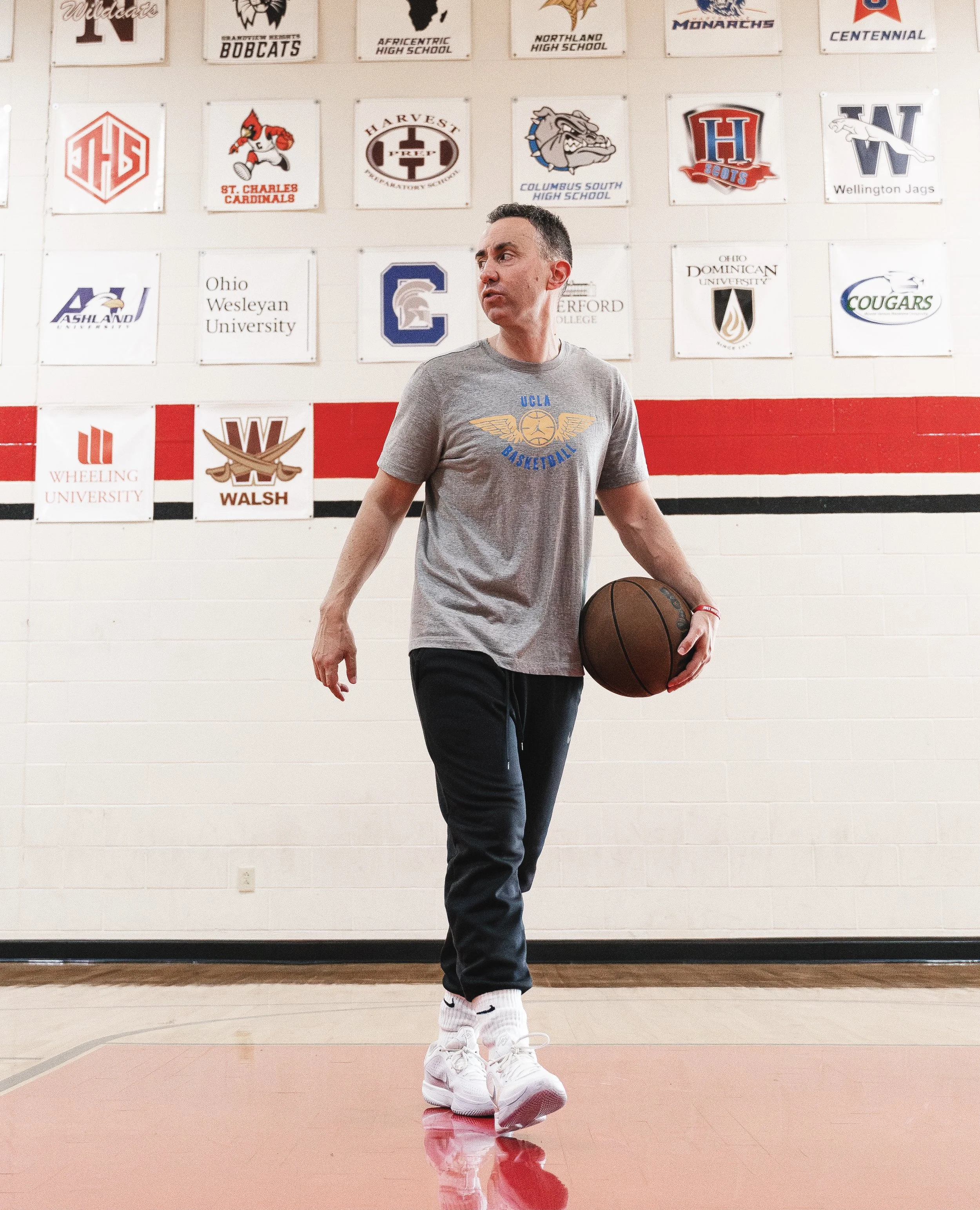 A man in a gray UCLA basketball t-shirt and black pants holding a basketball walking on a gymnasium court with school logos on the wall behind him.