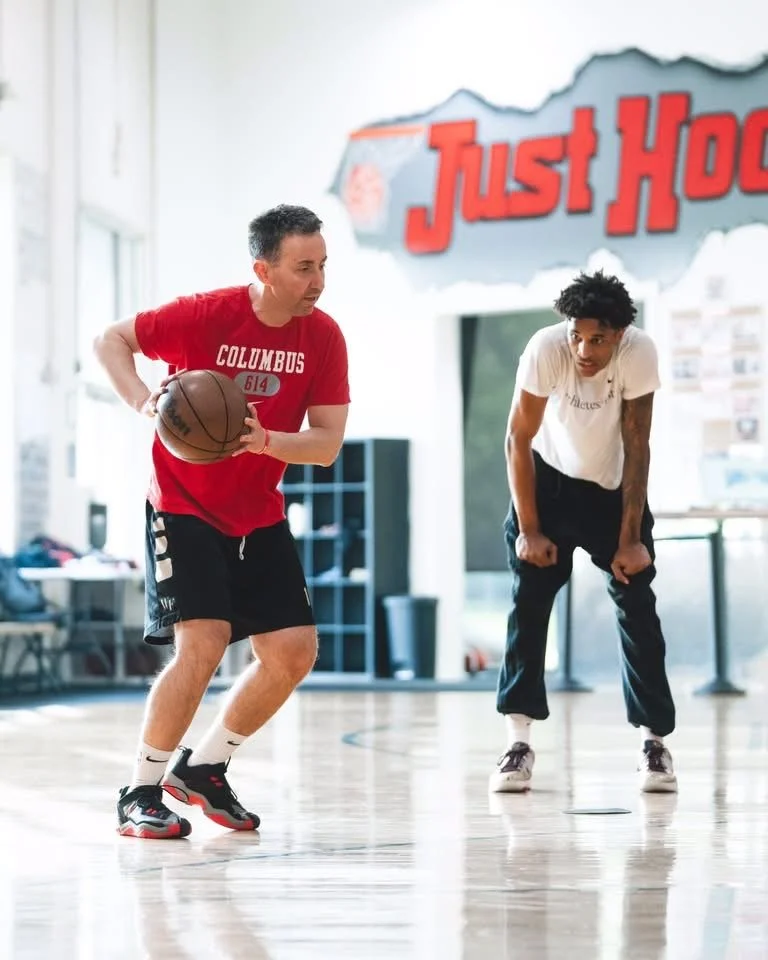Two men playing basketball in an indoor gym, with one holding a basketball and the other standing nearby, both focused on the game.