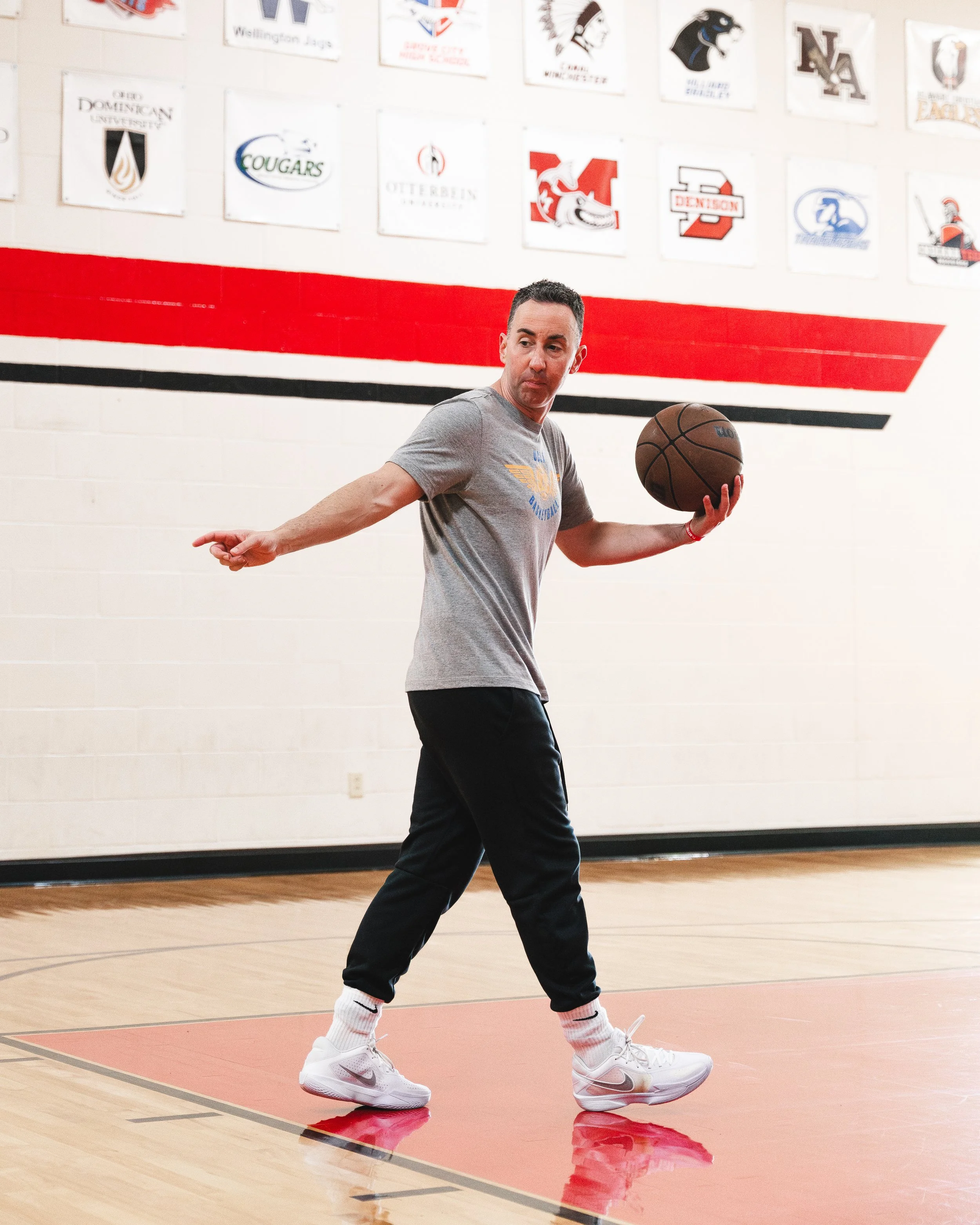 A man in gray t-shirt and black pants holding a basketball on an indoor basketball court, with sports banners on the wall behind him.