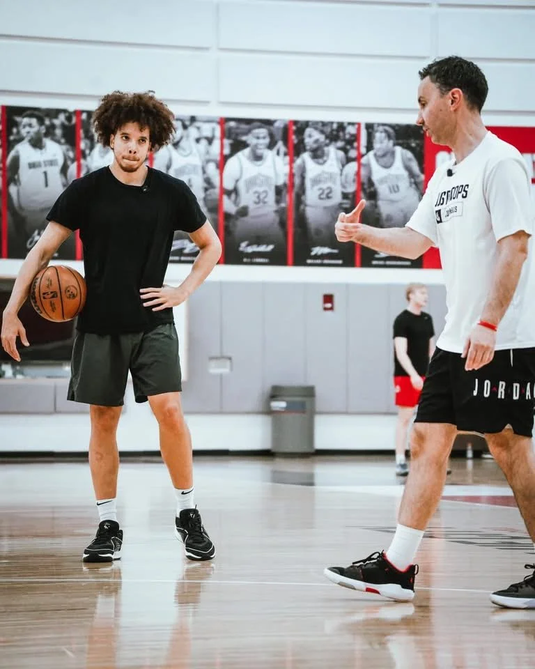 A young man with curly hair holding a basketball on a basketball court, listening to his coach who is giving instructions.
