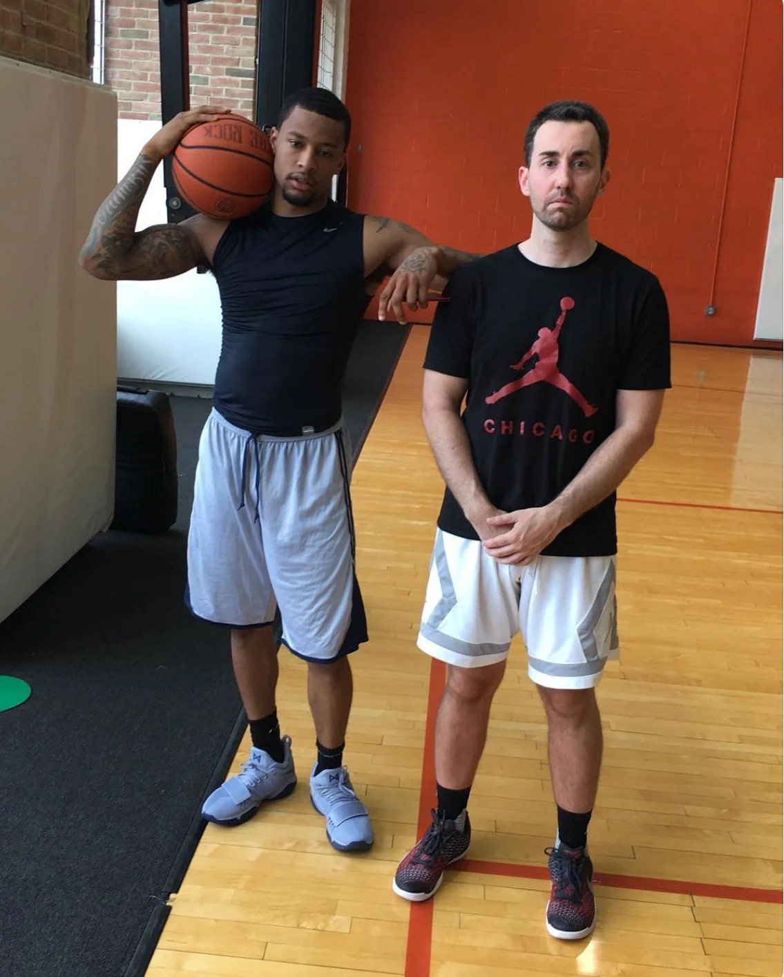 Two men standing on a basketball court. The man on the left is holding a basketball on his shoulder, wearing a black sleeveless shirt, shorts, and sneakers. The man on the right is wearing a black T-shirt with a red Jumpman logo and the word 'Chicago' underneath, shorts, and sneakers.