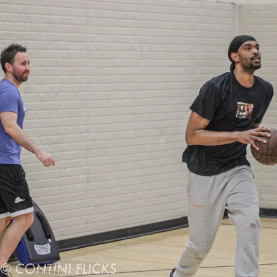 Two men playing basketball indoors, one dribbling the ball and the other watching him.