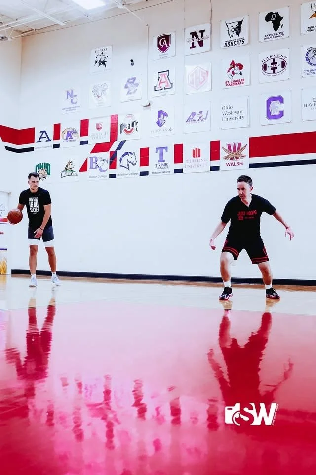 Two basketball players practicing on an indoor court with university banners on the wall behind them.