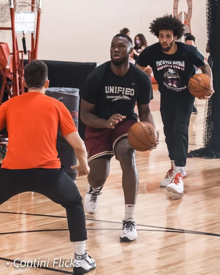 Two men playing basketball, one in a black shirt with 'UNIFIED' written on it, dribbling the ball, and another in a black T-shirt with a graphic, holding a basketball, in a gym with a wooden floor.