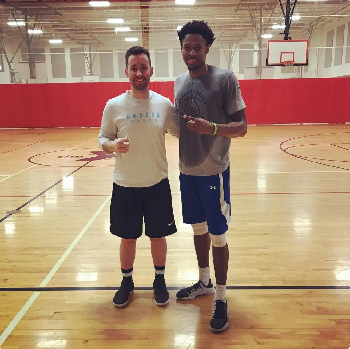 Two men stand together on an indoor basketball court, smiling and posing for a photo. One man wears a long-sleeve shirt and shorts, the other wears a t-shirt, shorts, and a knee brace.
