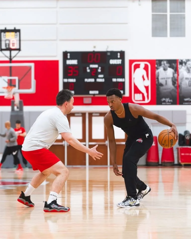 Two men playing basketball on an indoor court, one in a white shirt and red shorts defending, the other in a black tank top and black pants dribbling the basketball.