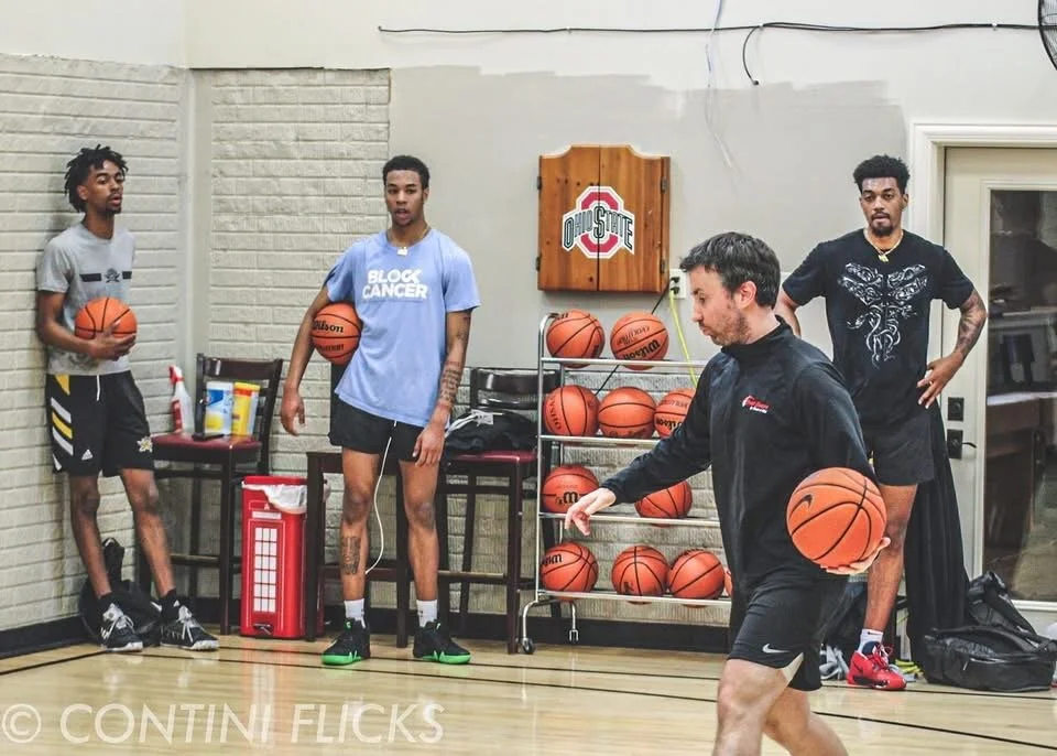 Four young men in a gym with basketballs, some holding basketballs, and a trainer practicing dribbling.