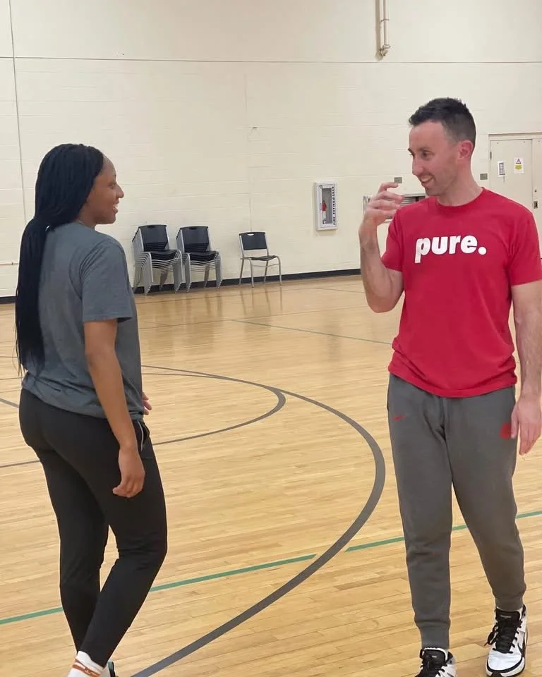 Two people in workout clothes smiling and talking inside a gymnasium with wooden flooring and empty chairs in the background.