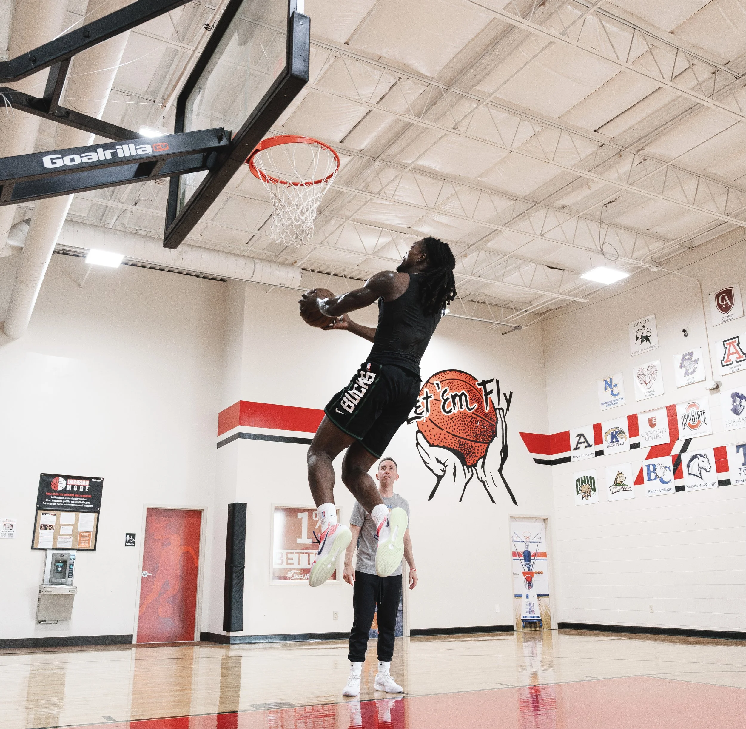 A person in mid-air playing basketball in a gym, with another person watching beneath them. The gym has a basketball hoop and banners on the wall.