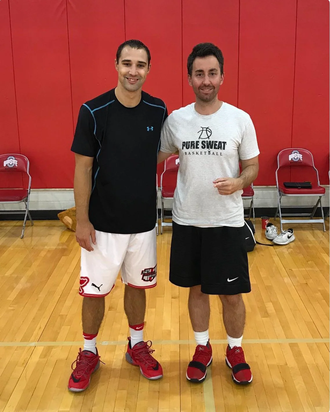 Two men standing on an indoor basketball court, smiling at the camera. They are dressed in athletic clothing, with one wearing a black undershirt and white shorts and the other in a grey t-shirt and black shorts. Behind them are red chairs and red wall panels.