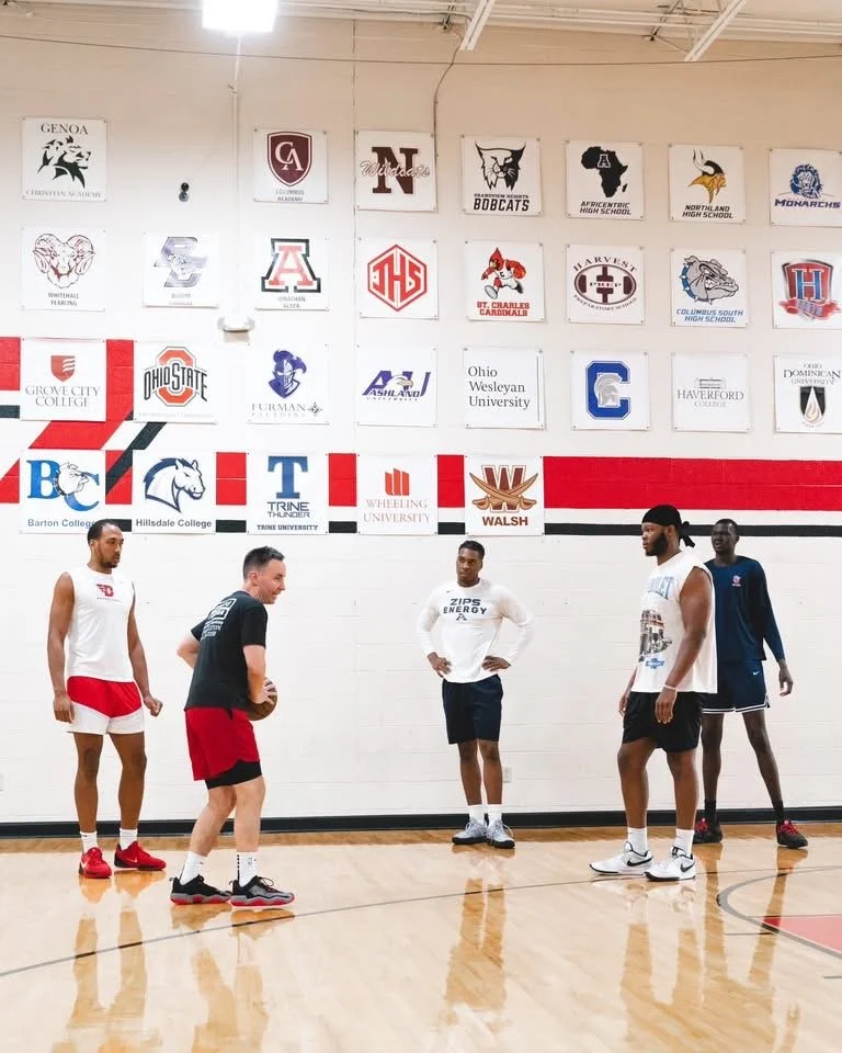 Five basketball players standing on a gym court, one holding a basketball, with college banners hanging on the wall behind them.