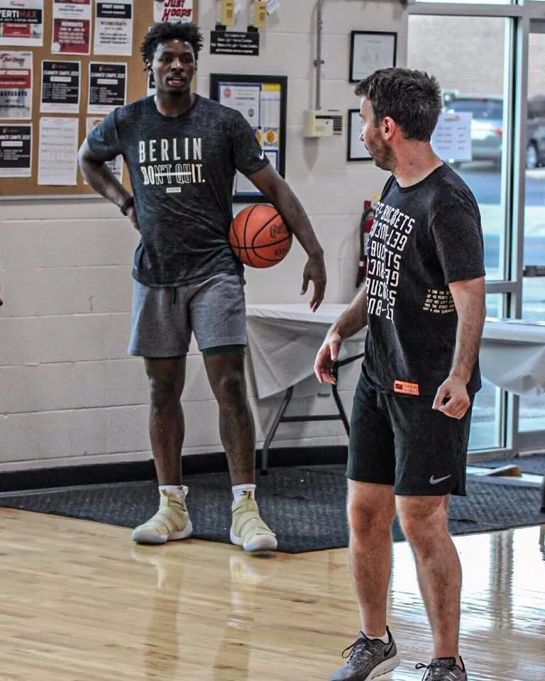 Two men in athletic clothing talking inside a gym; one holding a basketball, the other facing him, near a bulletin board and large windows.