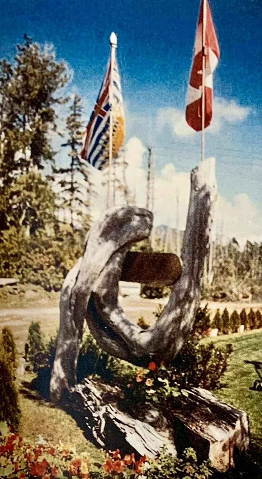 A large wooden entrance sign at Mackenzie Beach Resort, situated in a well-maintained garden with flowers, with two flags in the background, one British Columbian and one Canadian. There are trees and buildings visible behind the flags.
