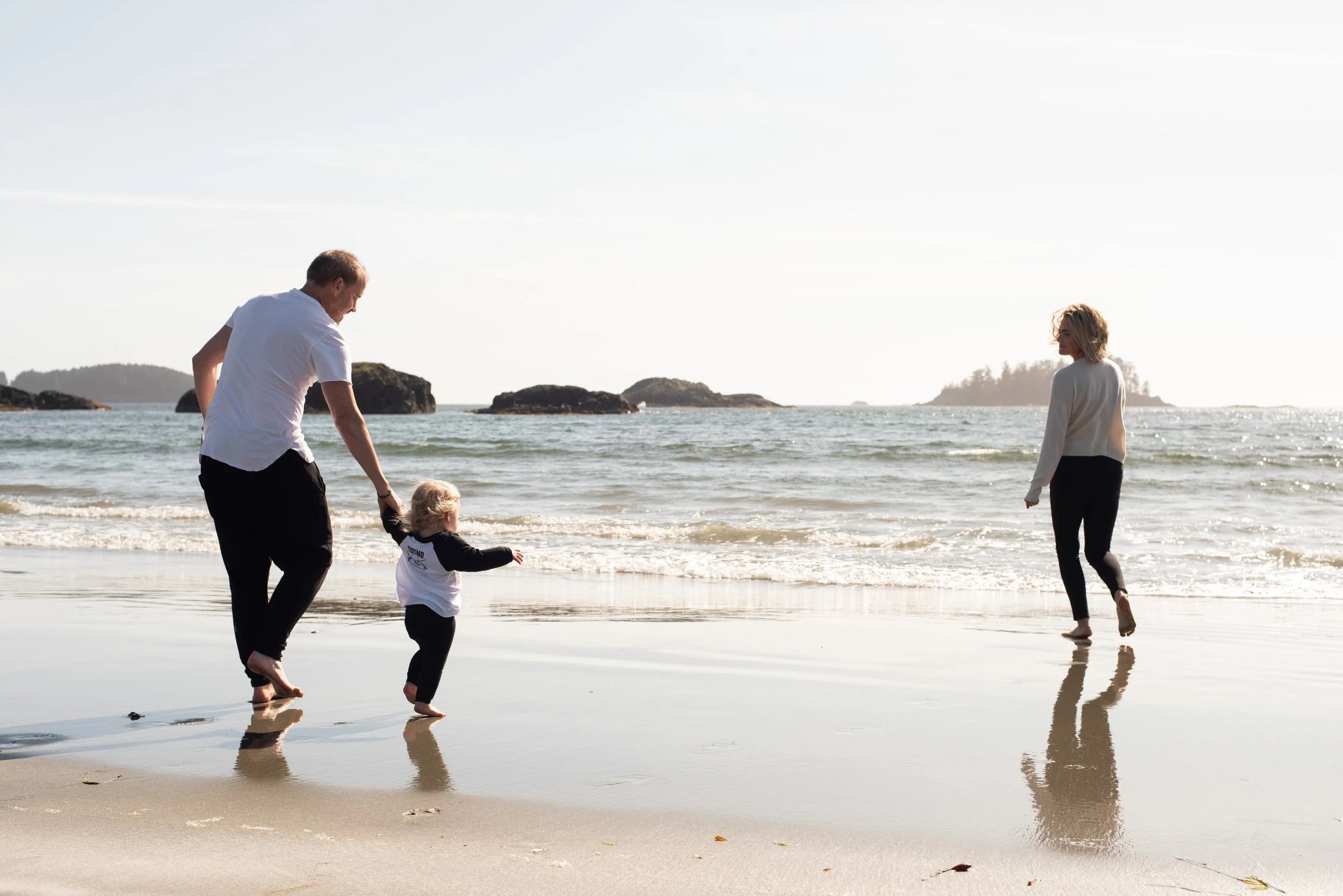 A family walking along the shoreline on a beach with rocks in the water, a man holding a child's hand, and a woman walking ahead.