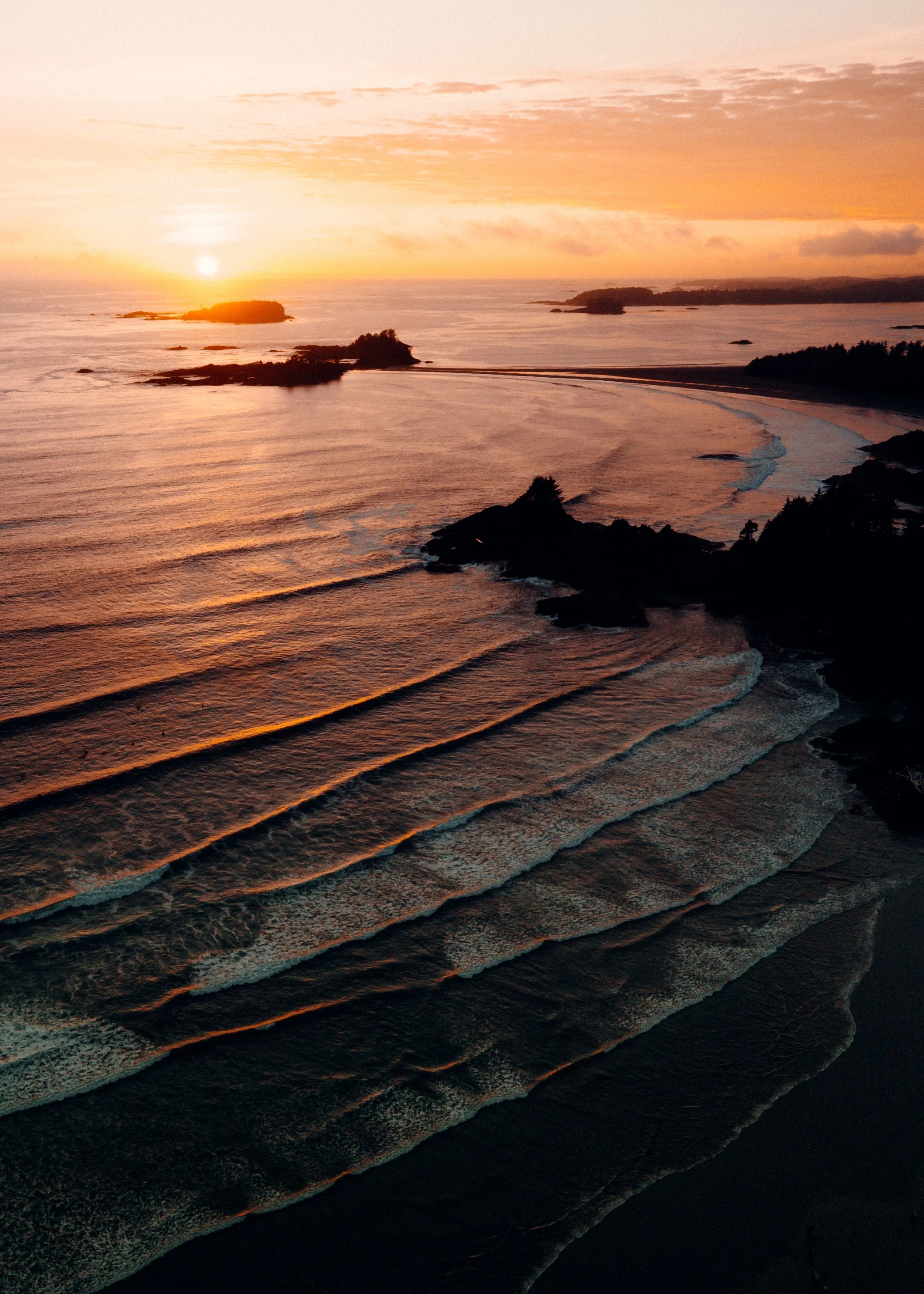 Sunset over a coast with calm waves, rocky islands, and a sandy beach, viewed from above.
