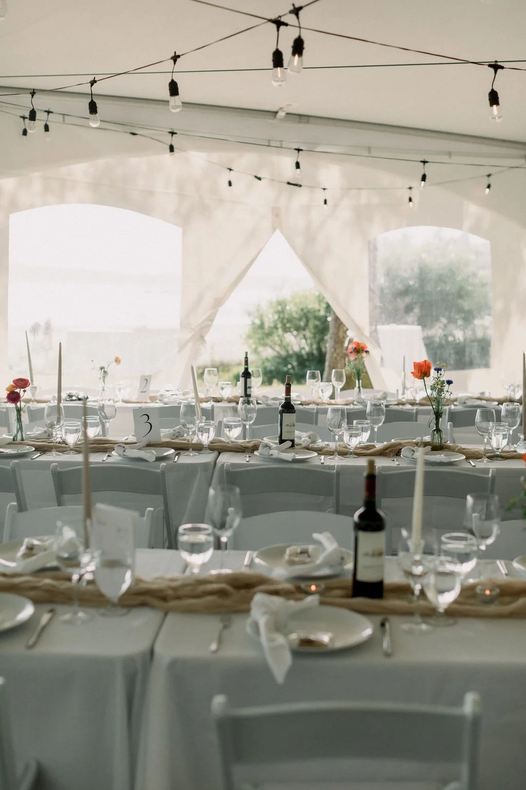 Elegant wedding or event reception setup inside a white tent with draped fabric and string lights. Long tables are decorated with flowers, wine bottles, glasses, candles, and table number cards.