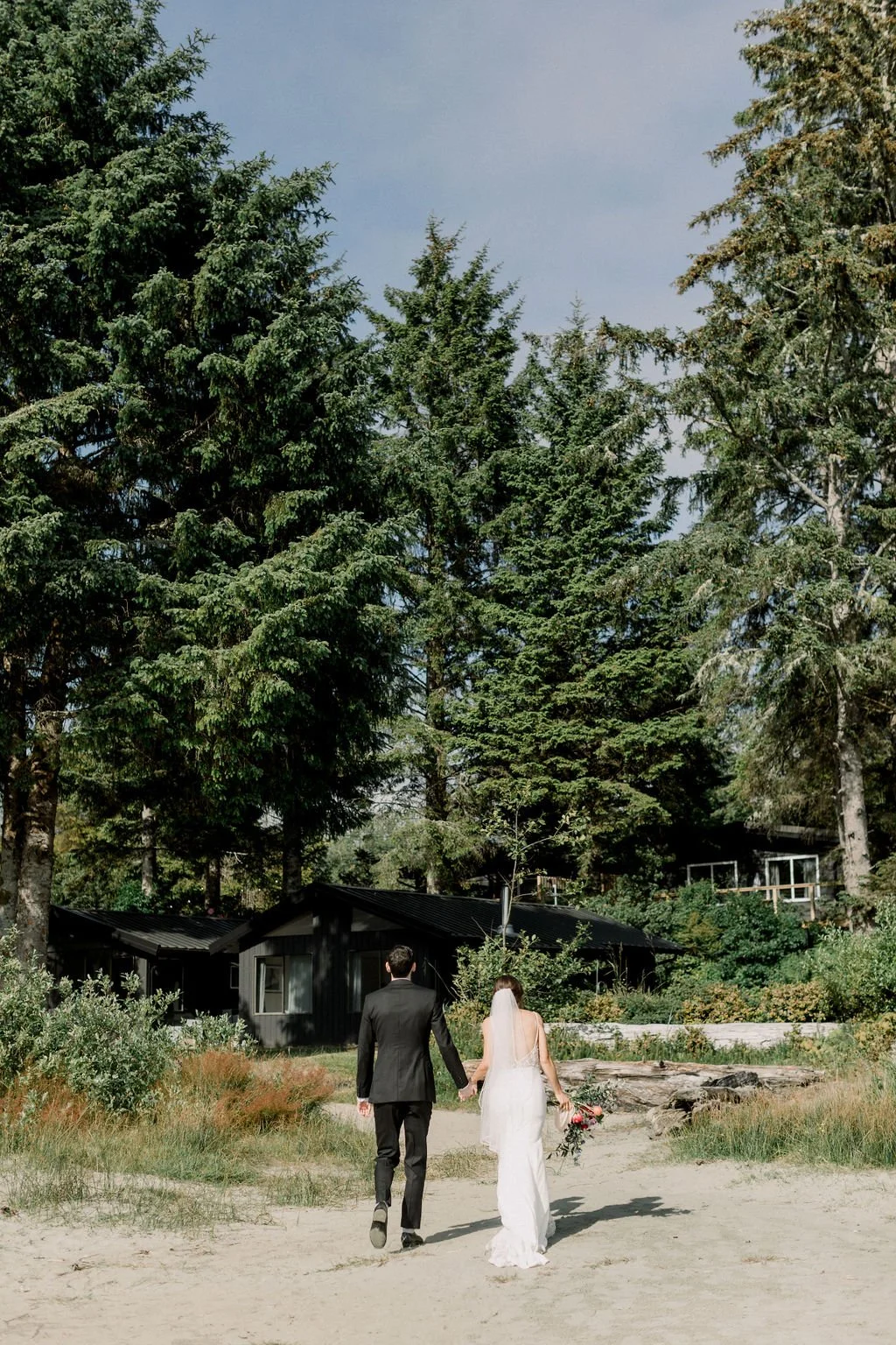 A bride and groom walking hand in hand on a sandy path through a lush, green outdoor area surrounded by tall pine trees, with black cabins in the background and a cloudy sky overhead.