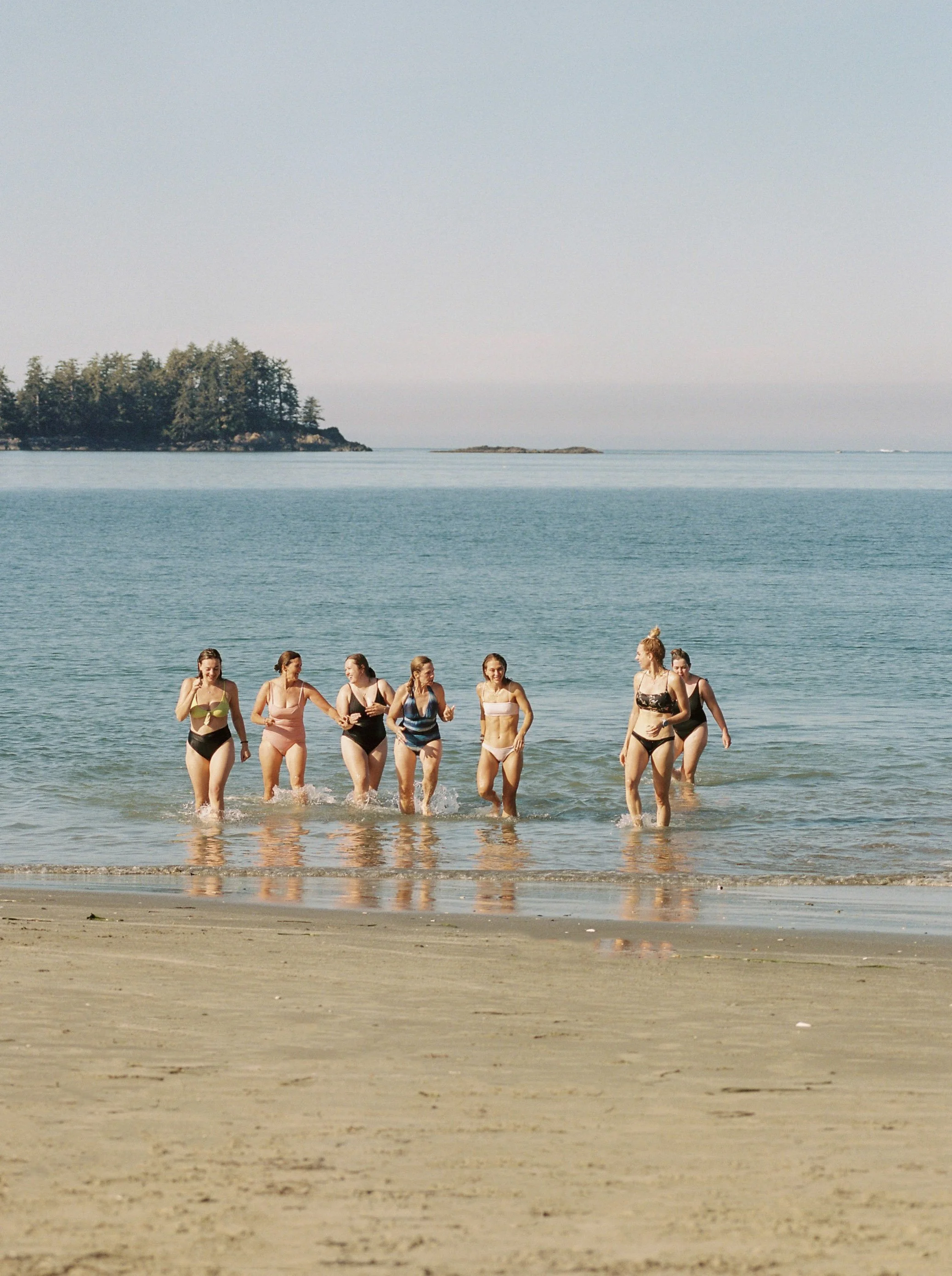 Group of women in swimsuits walking out of the ocean onto the sandy beach, with trees and small islands in the background.