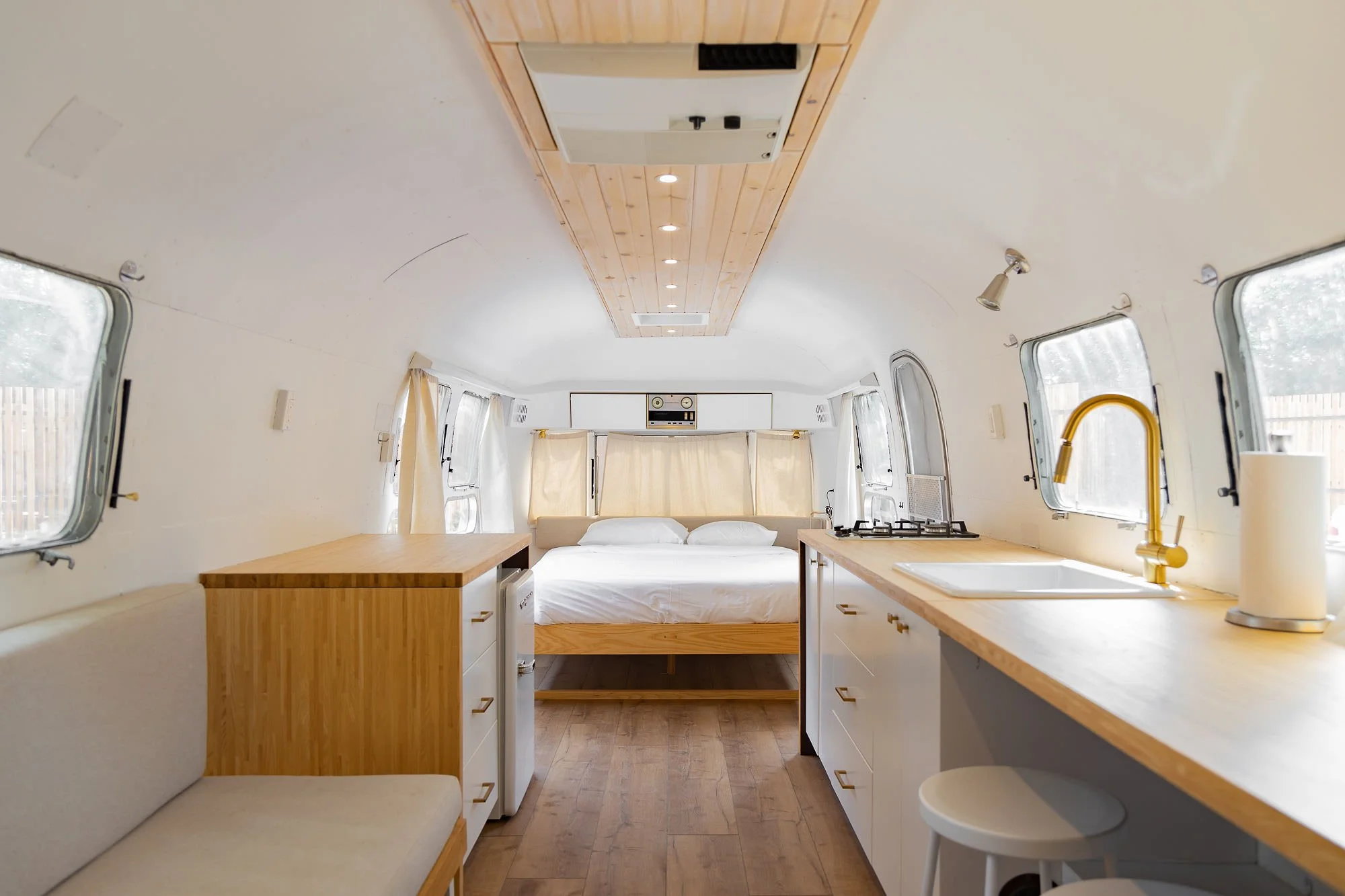 Interior of a small, cozy camper with a sleeping area, kitchen, and seating area, featuring white walls, wooden accents, and multiple windows.