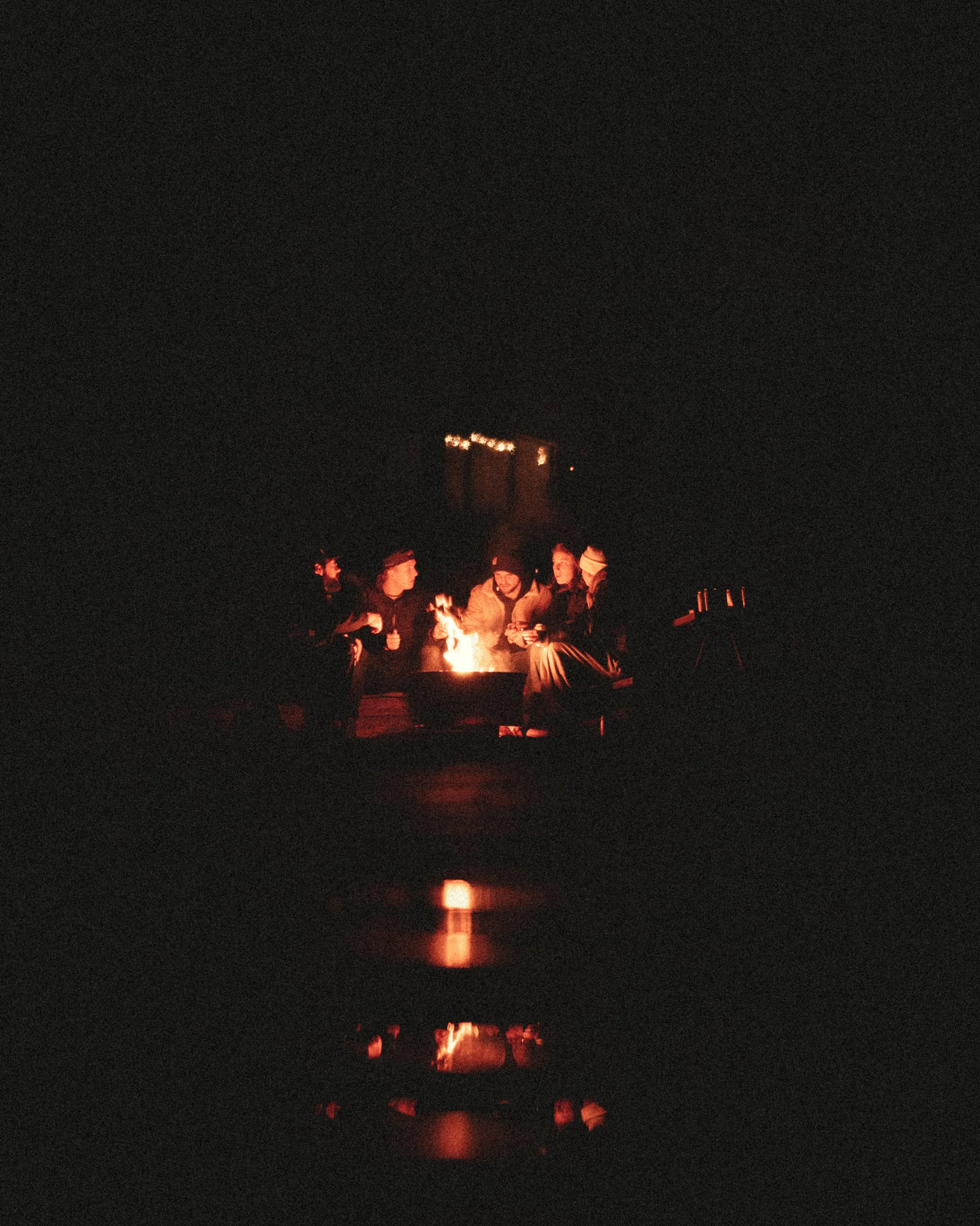 A group of people sitting around a campfire at night, with a dark sky and their reflections visible on water.