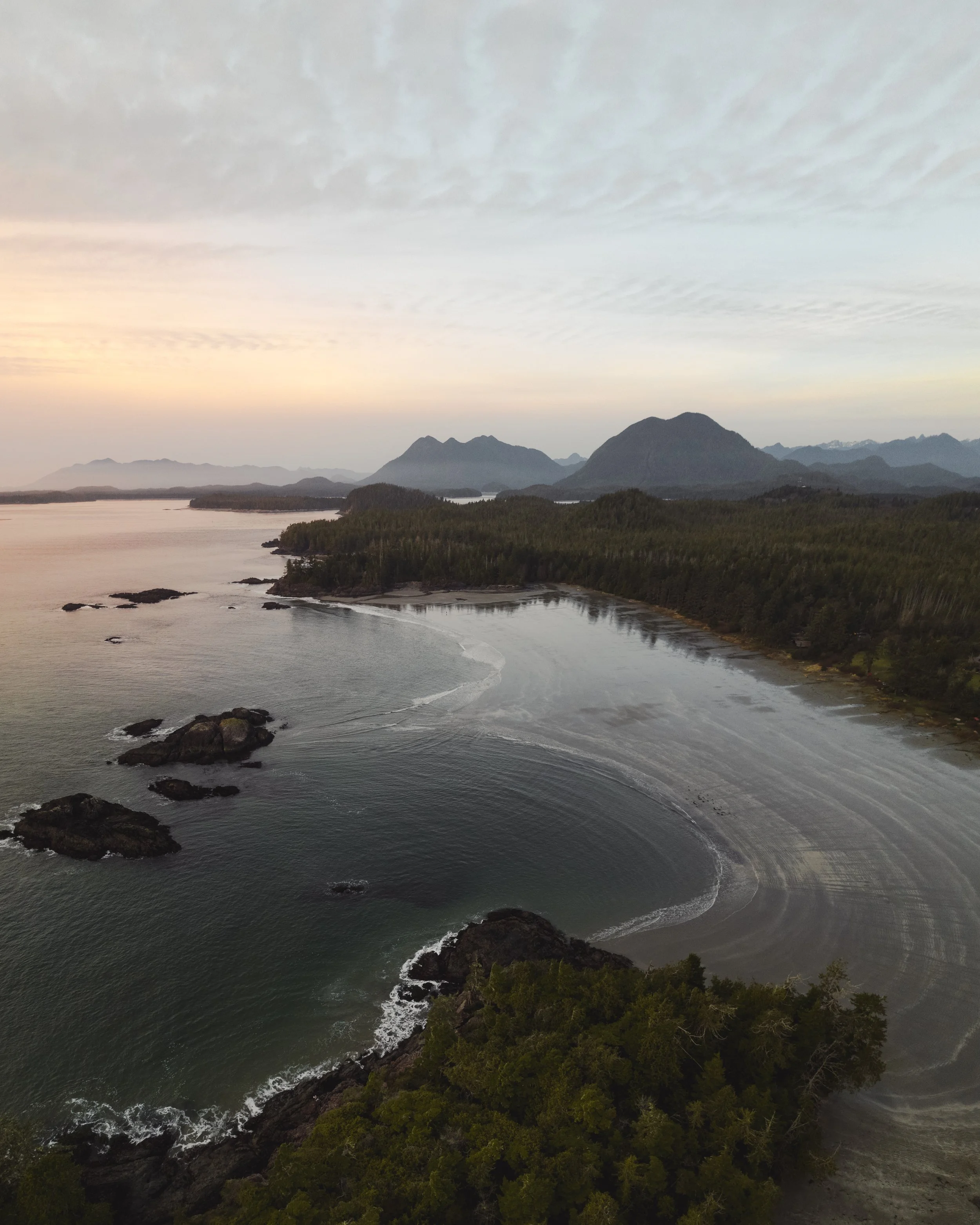 Aerial view of rocky shoreline, surf, and dense forest with mountain range in background at sunset.