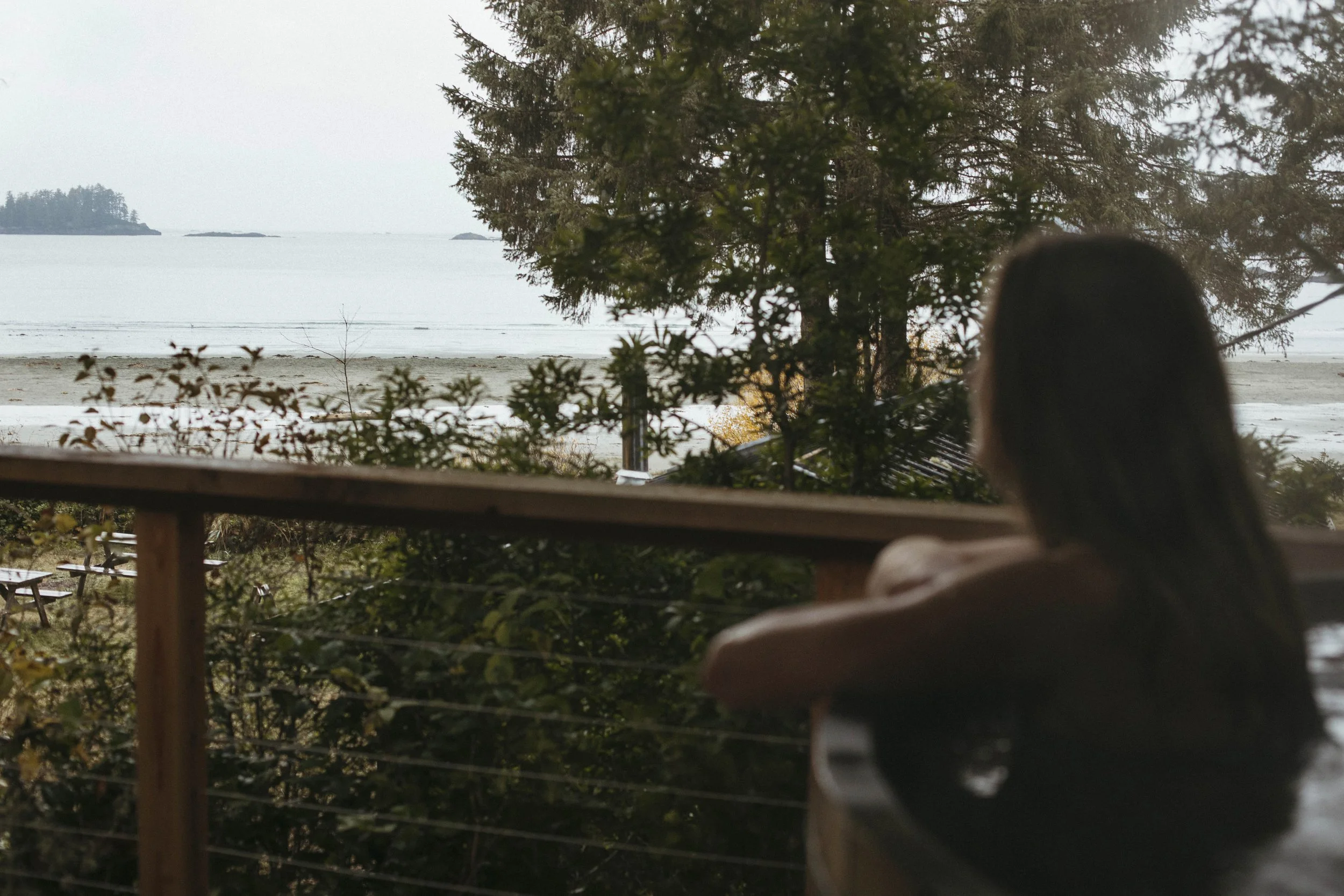 A person with long dark hair sitting on a balcony overlooking a beach with trees and water in the distance.