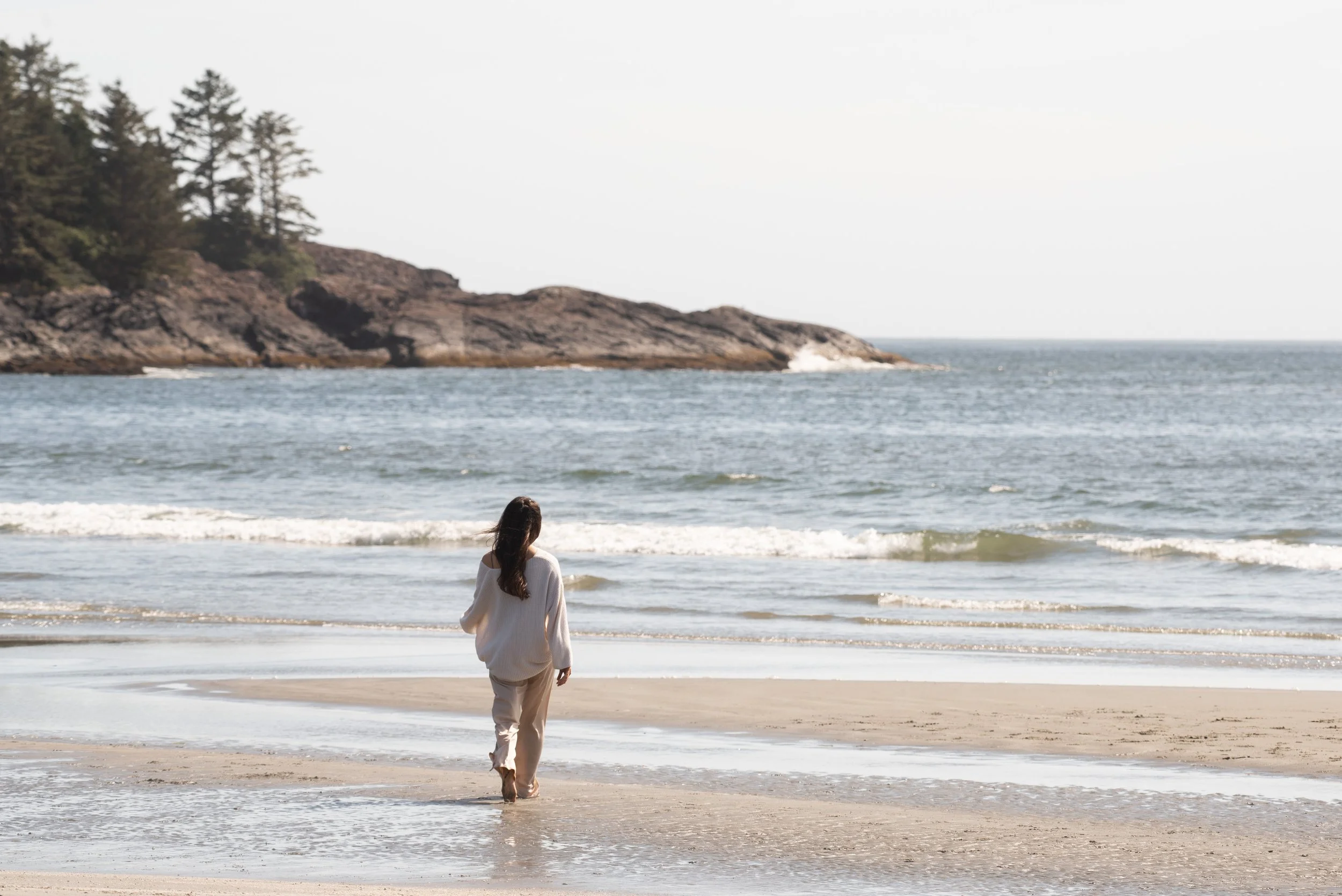 A woman walking barefoot on a sandy beach near the water's edge with waves, greenery-covered rocky cliffs, and an overcast sky in the background.