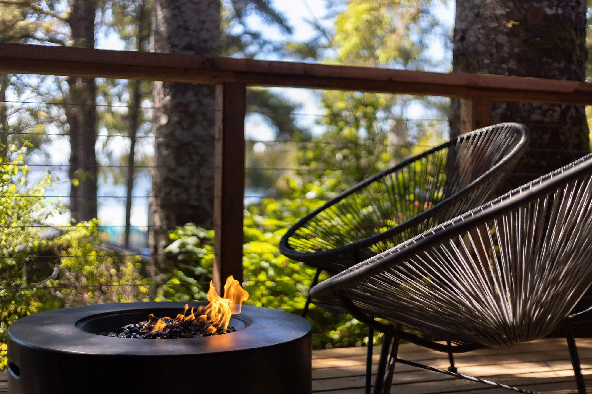 Outdoor deck with two black wire chairs and a fire pit with a small flame, surrounded by trees and green foliage.