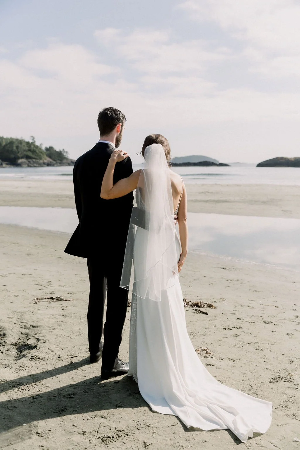 A bride and groom standing on a beach, with the bride in a white wedding gown and veil, and the groom in a black suit, facing the ocean during their wedding photoshoot.