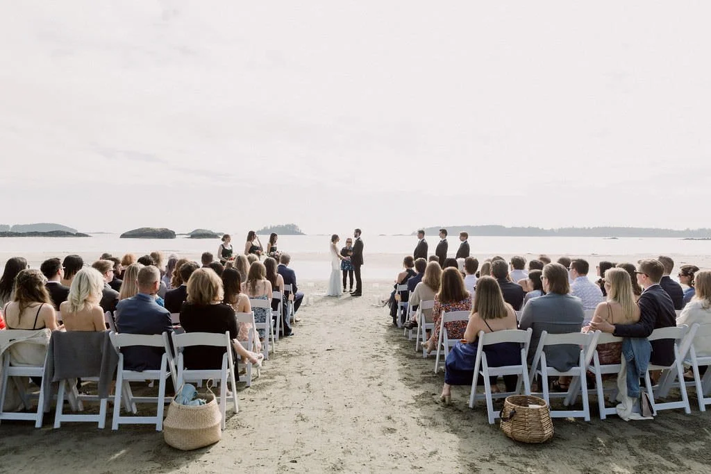 A wedding ceremony on a beach with guests seated in white chairs, the couple exchanging vows at the altar, and the ocean in the background.