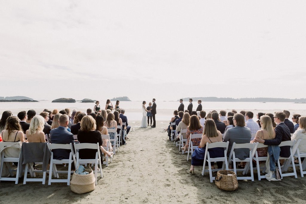 A beach wedding ceremony with guests seated on white chairs facing the ocean, where a couple is getting married with an officiant and bridesmaids and groomsmen standing nearby.