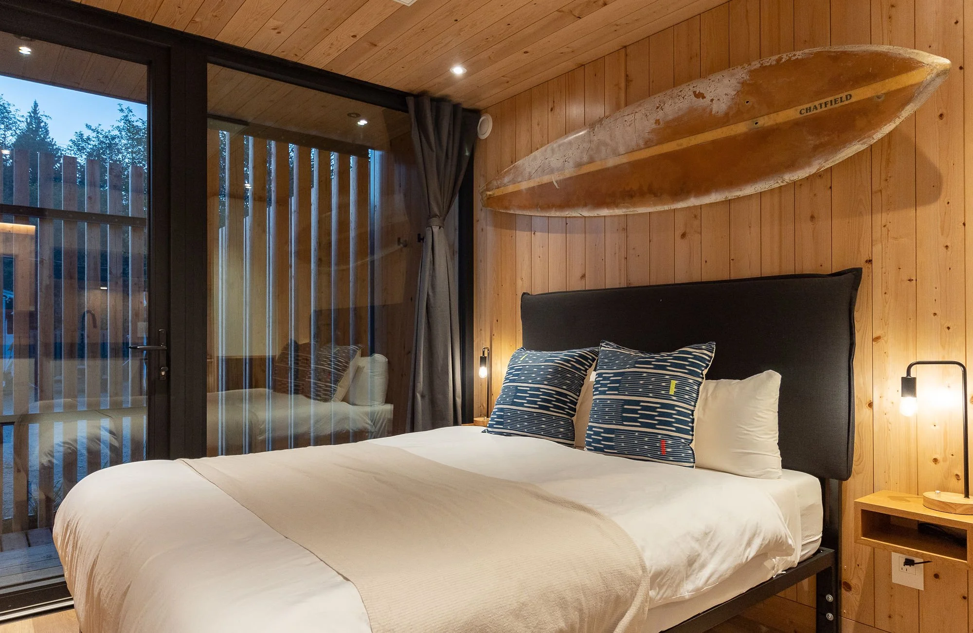 Cozy bedroom with wooden walls and ceiling, featuring a large bed with blue patterned pillows and a black headboard. A surfboard decor hangs above the bed. Large glass sliding door shows a partial view of a balcony with outdoor seating and trees outside.