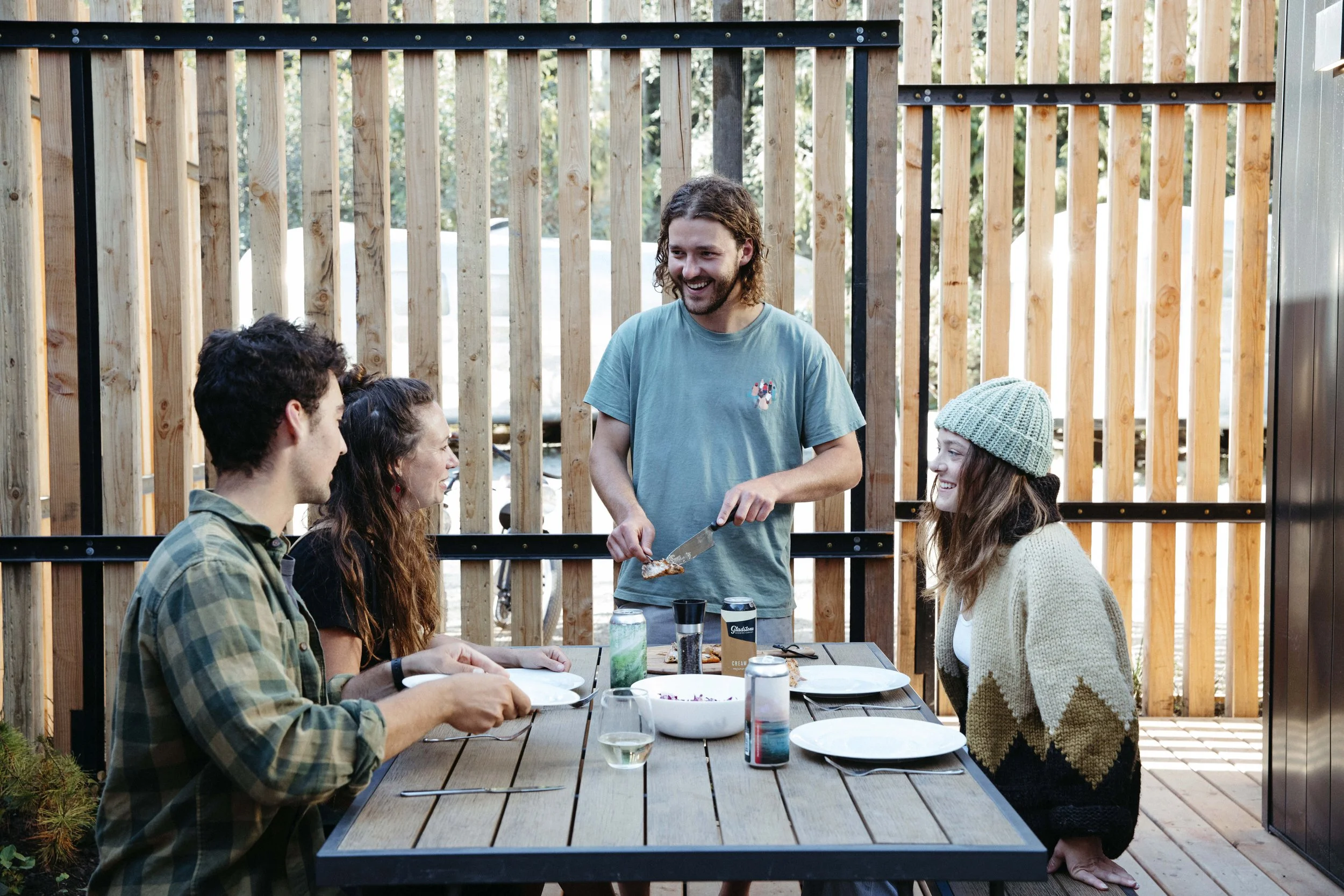 Four young adults enjoying a backyard barbecue, with one man serving food while the other three sit at a table with plates and drinks in front. They are in front of a wooden fence and trees, and the mood appears cheerful and relaxed.
