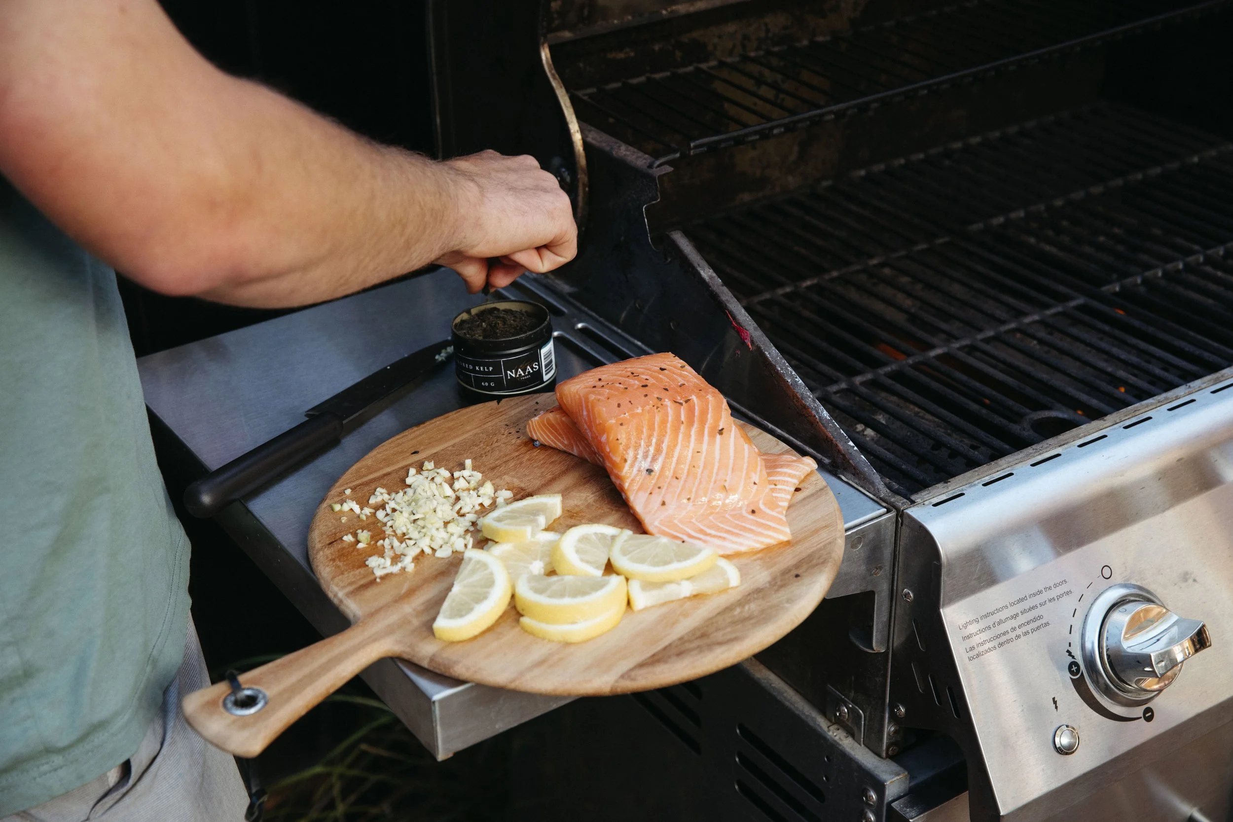 Person seasoning salmon fillet on a grill with lemon slices, minced garlic, and black pepper.
