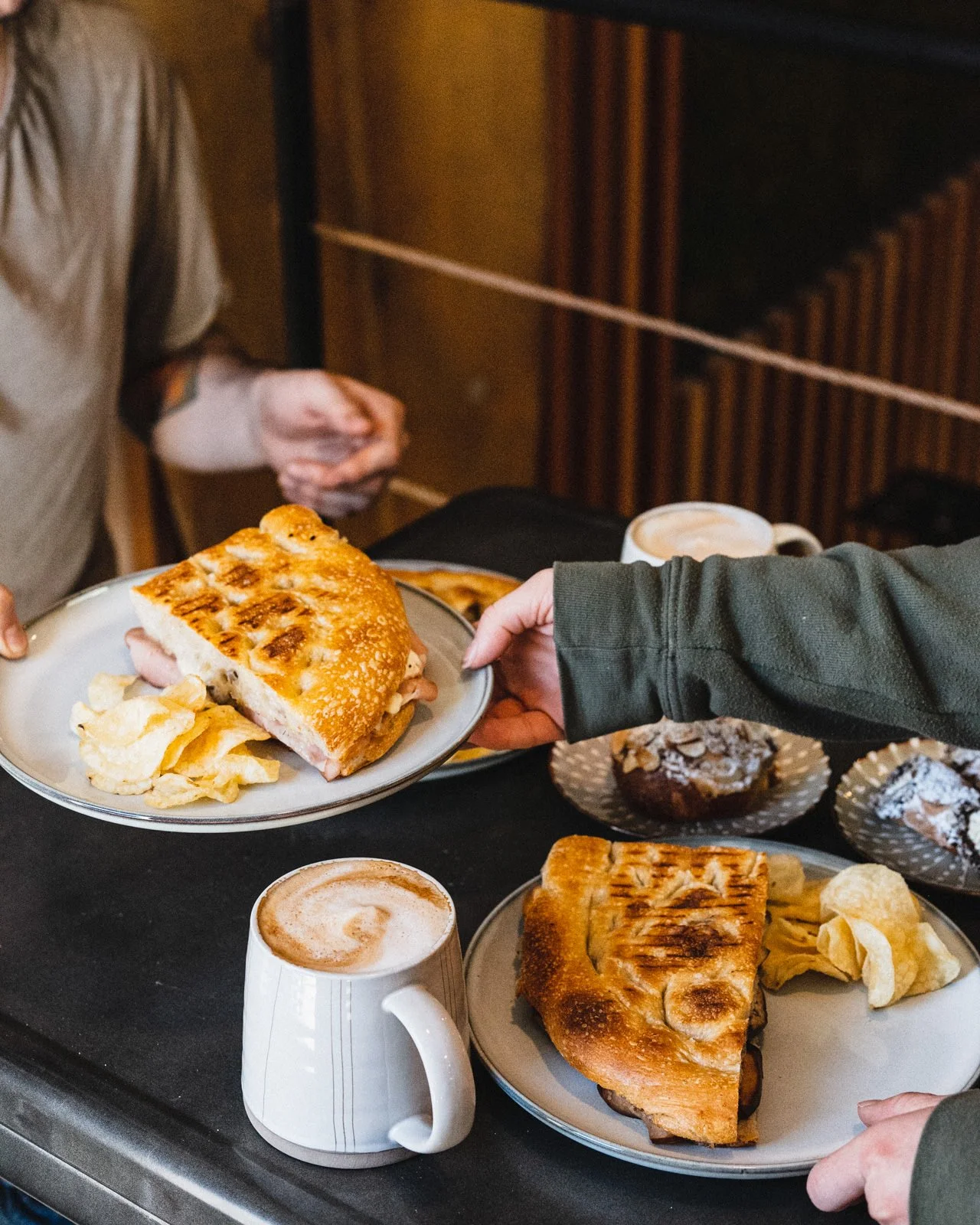 Two plates of breakfast sandwiches with potato chips, a cup of coffee, and muffins on a black table.